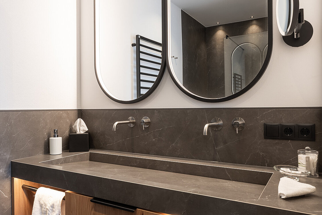 Modernes Badezimmer mit Doppelwaschbecken, zwei großen ovalen Spiegeln und einem Holzunterschrank. Handtücher sind vorhanden, und eine diskrete Beleuchtung schafft eine elegante Atmosphäre.