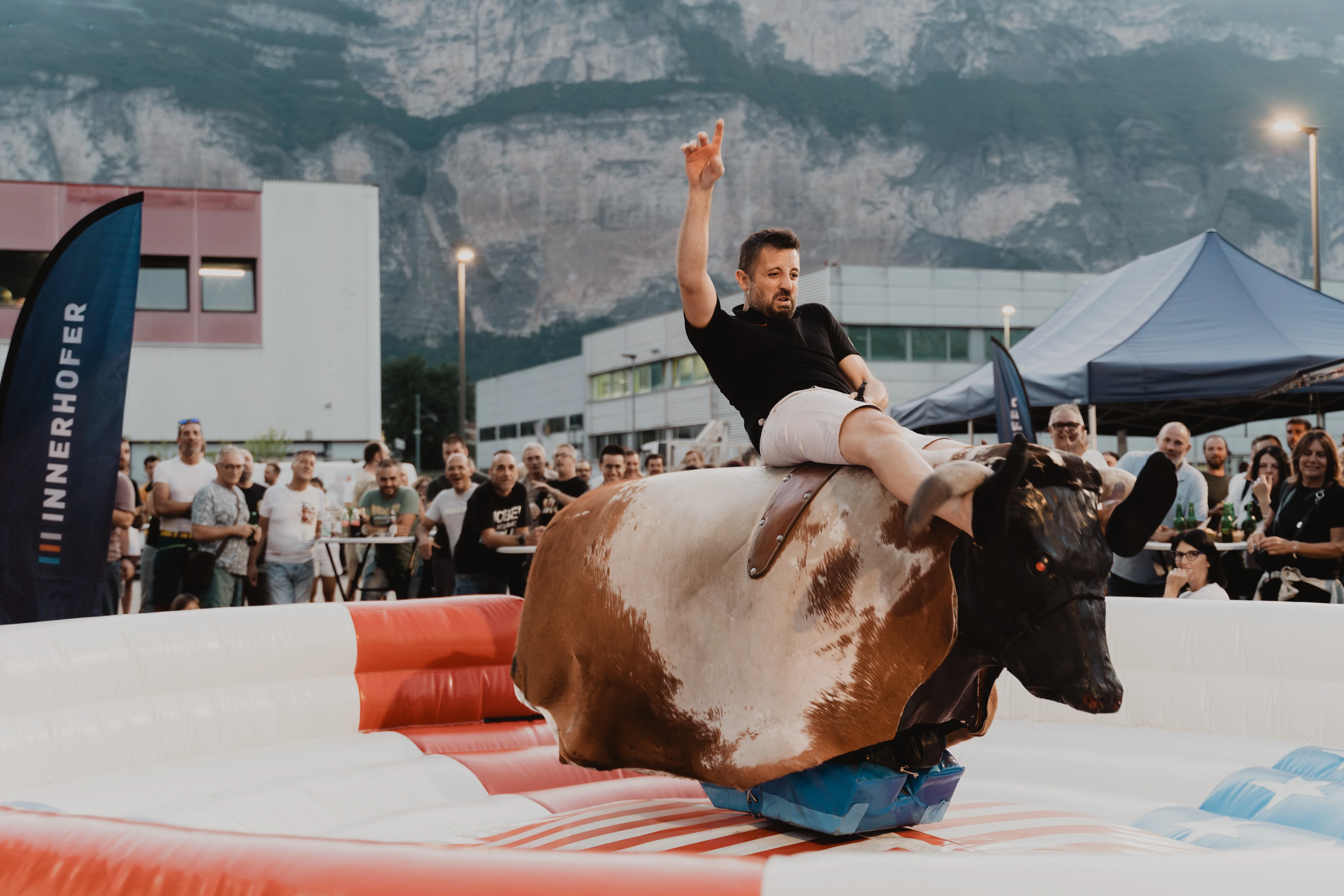 Ein Mann sitzt auf einem mechanischen Bullen bei einer Outdoor-Veranstaltung. Im Hintergrund stehen Zuschauer vor einem grauen Gebäude und einer Berglandschaft. Der Mann hebt eine Hand siegreich in die Luft, während er den Bullen reitet. Die Stimmung ist ausgelassen.
