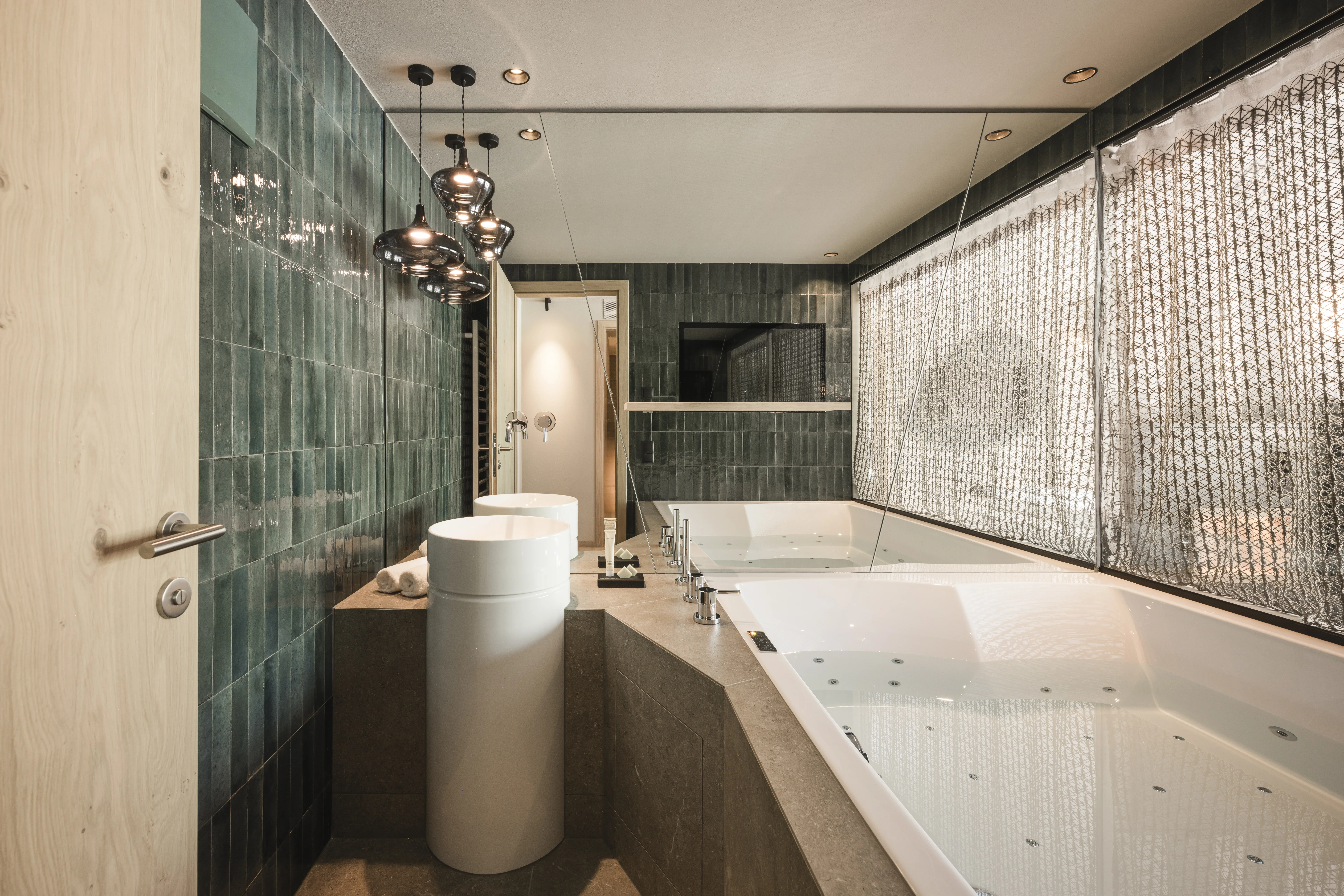 Ein modernes Badezimmer mit grünen Fliesen, einem großen Schiebefenster mit Vorhang, einer großen weißen Badewanne und einer freistehenden Waschbecken-Säule. Über dem Waschbecken hängen stilvolle Lampen. Ein Spiegel erstreckt sich über die gesamte Wand über der Badewanne.