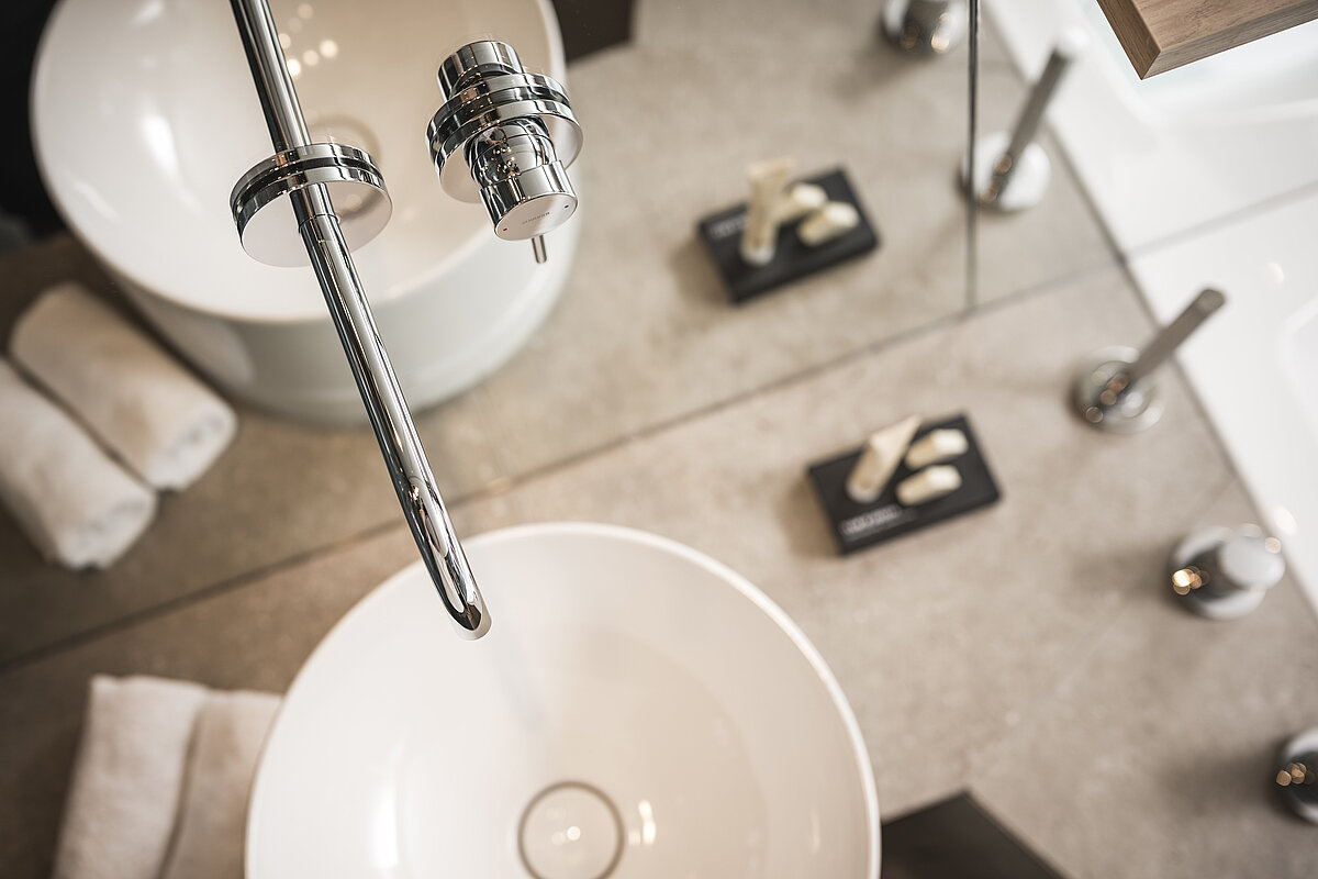 Modernes Badezimmer mit stilvollem, kreisförmigem Waschbecken, Chromarmaturen und elegant platzierten Handtüchern. Daneben liegen auf dunklen Unterlagen kleine Fläschchen mit Pflegeprodukten.