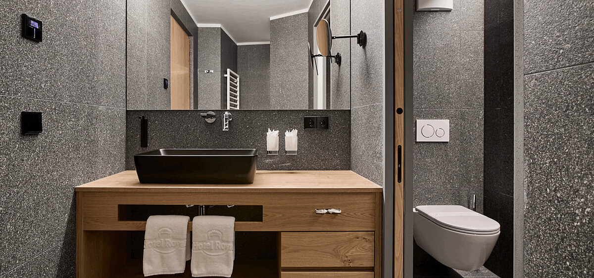 Modernes Badezimmer mit dunkler Steinwand, großem Spiegel, Holzschränken und Badewanne. Im Hintergrund ist ein Flur mit Tageslicht zu sehen.