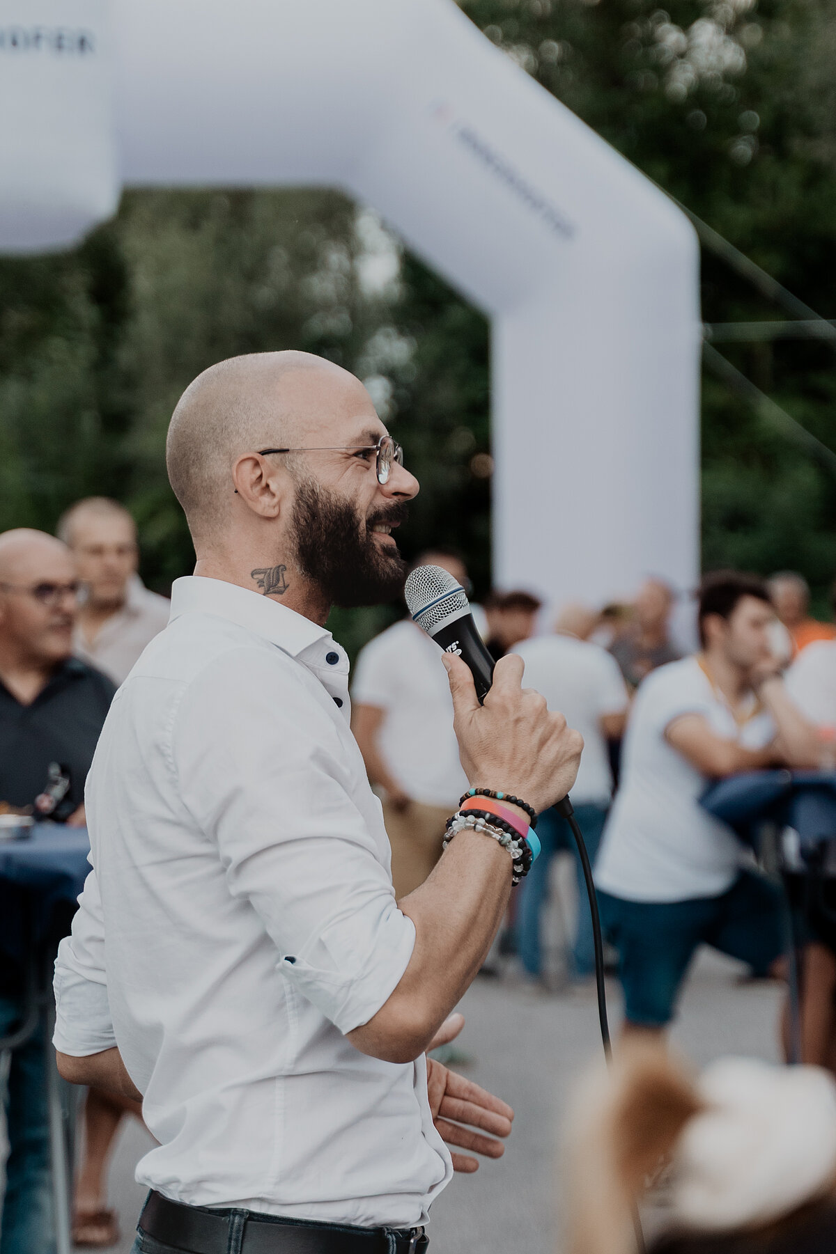 Ein Mann mit Brille und Mikrofon spricht vor einer Gruppe von Menschen im Freien. Er trägt ein weißes Hemd und hat ein Tattoo am Hals. Im Hintergrund sieht man eine aufblasbare Torbogenstruktur.