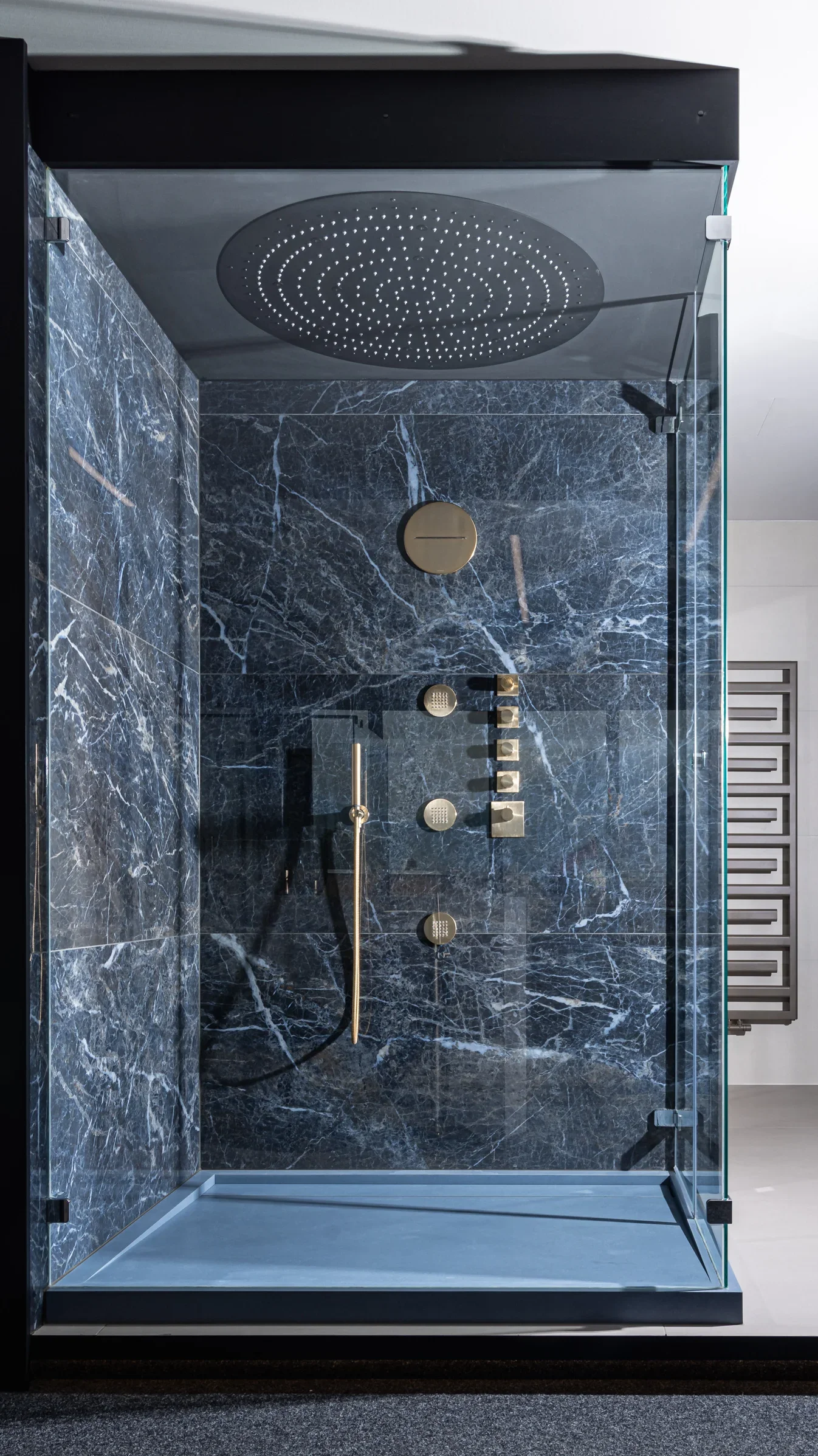 Luxuriöse Duschkabine aus Glas mit schwarzer Marmorverkleidung, wandintegrierten Armaturen und einem großen, runden Regenduschkopf an der Decke. Die Armaturen sind in Goldtönen gehalten. Im Hintergrund befindet sich ein moderner Handtuchhalter.