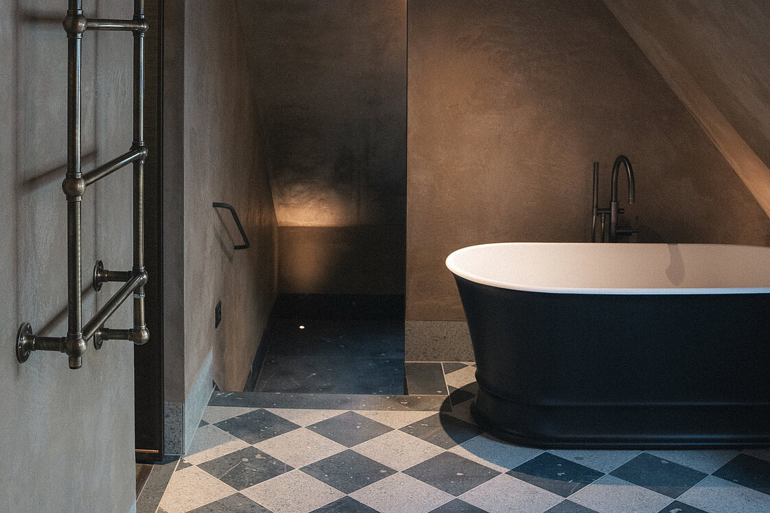 Ein modernes Badezimmer mit schräger Decke zeigt eine freistehende, dunkle Badewanne vor einer sanft beleuchteten Wand. Der Boden hat ein schwarz-weißes Schachbrettmuster. An der Wand links hängt ein verchromter Handtuchhalter. Das Ambiente ist ruhig und stilvoll.