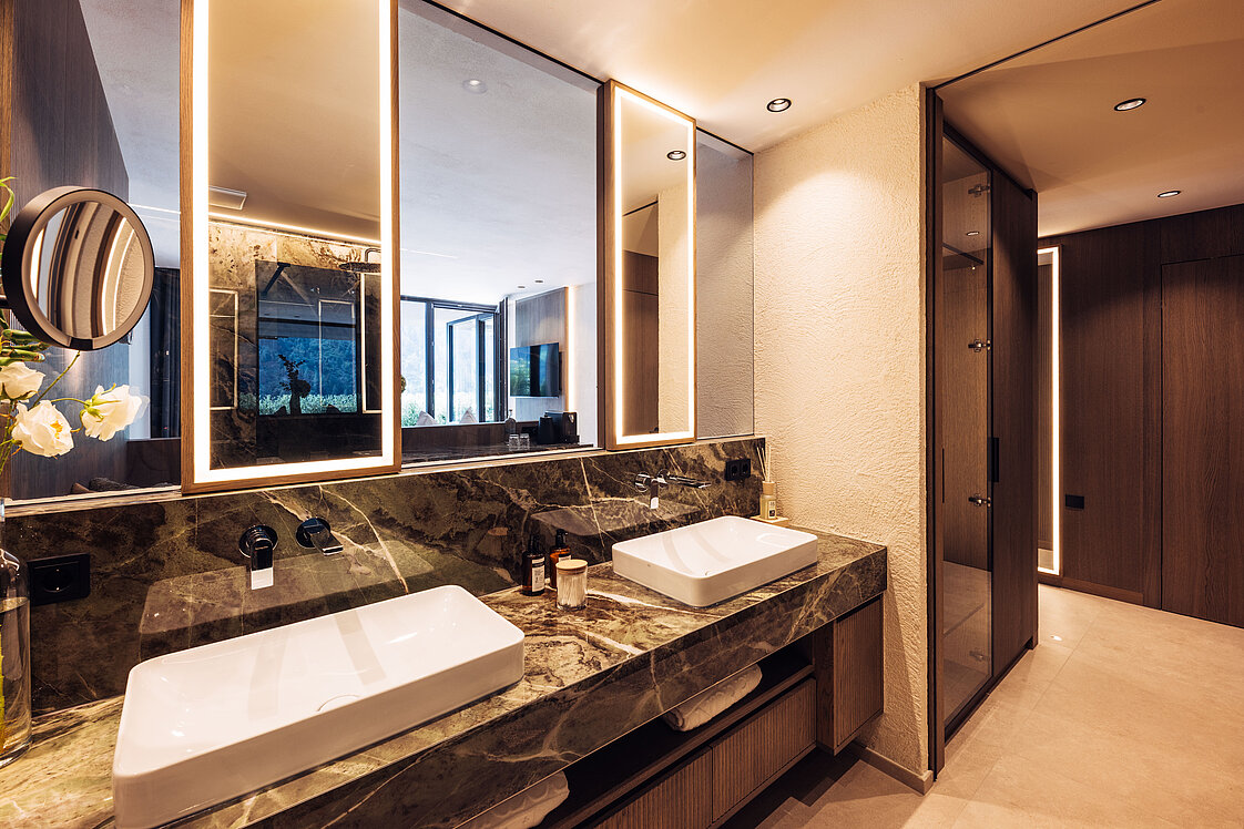 Modernes Badezimmer mit zwei rechteckigen Waschbecken auf einer eleganten Marmorablage. Große, hinterleuchtete Spiegel hängen darüber. Der Raum ist in warmen Farbtönen gehalten, mit Holzverkleidungen und diskreter Beleuchtung. Im Hintergrund ist ein Wandbildschirm zu erkennen.