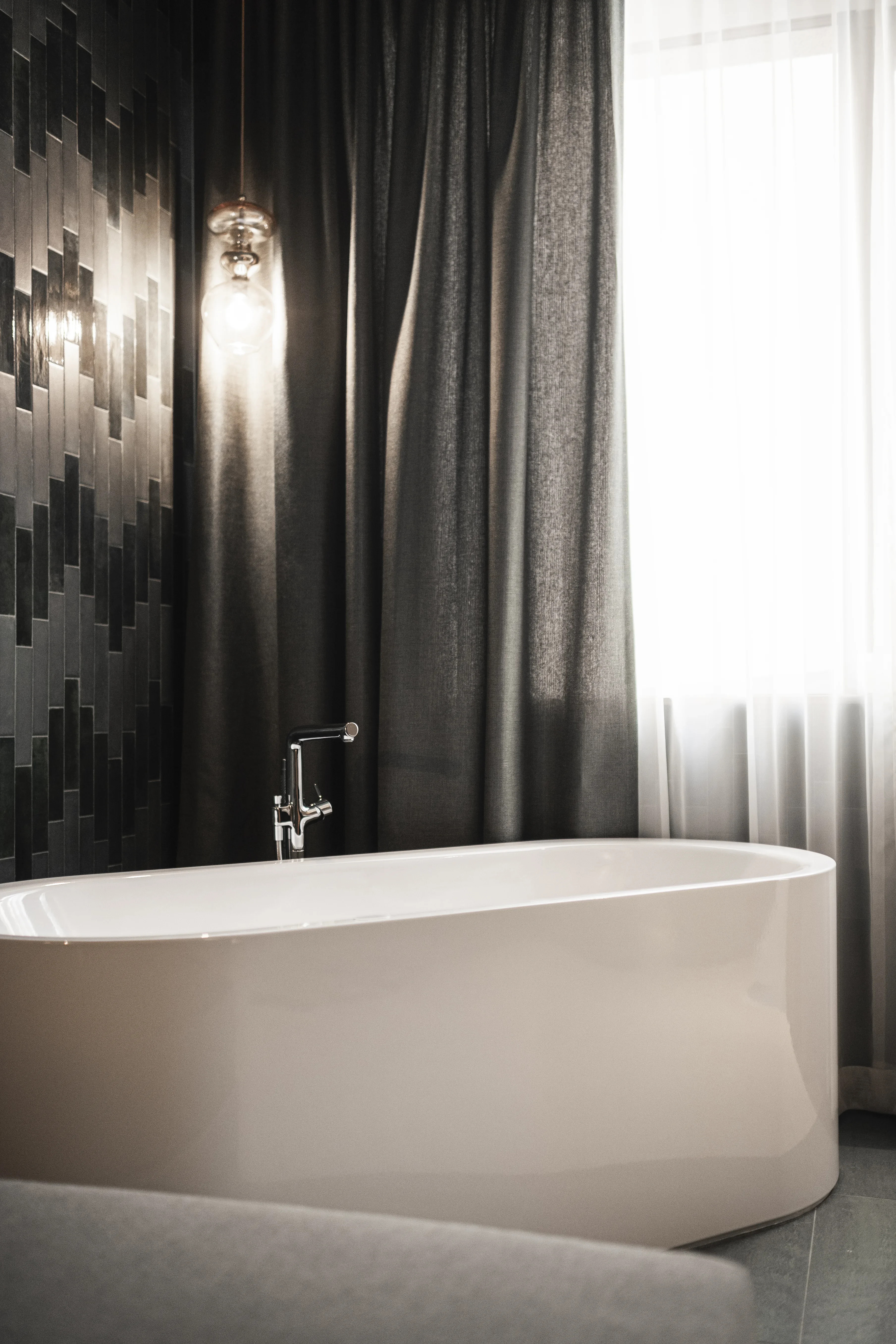 Moderner Badezimmerbereich mit einer freistehenden weißen Badewanne vor einem gemusterten dunklen Fliesenhintergrund. In der Nähe hängen Vorhänge, die teilweise ein Fenster verdecken, durch das weiches Licht fällt. Eine hängende Glühlampe beleuchtet die Szene sanft.