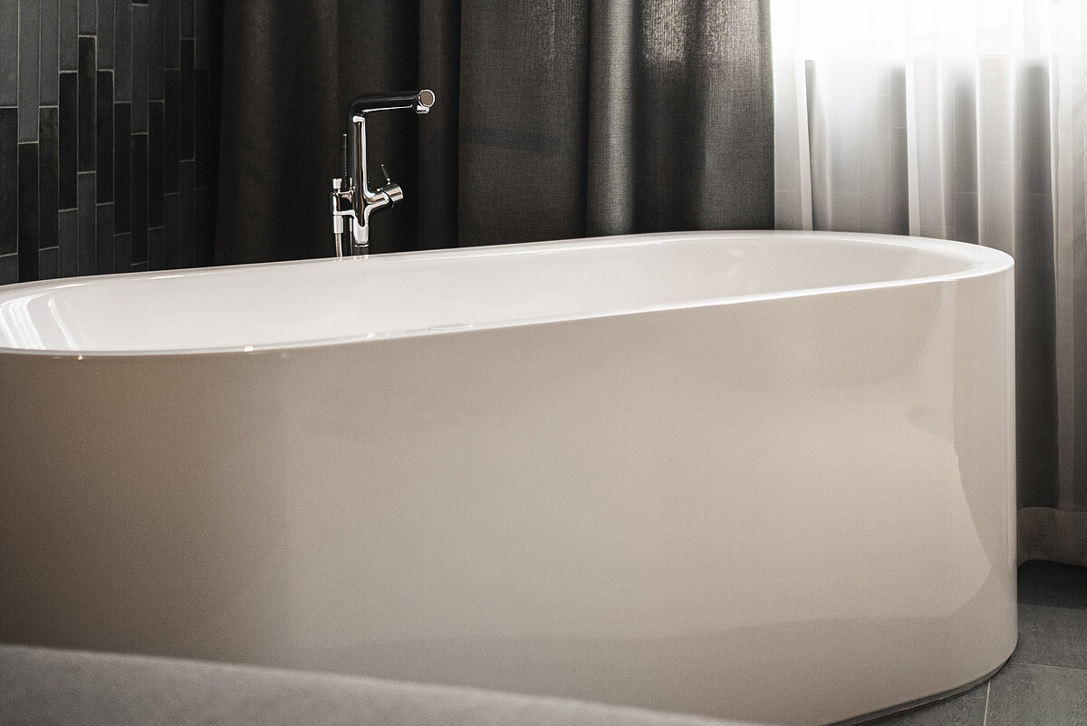 Moderner Badezimmerbereich mit einer freistehenden weißen Badewanne vor einem gemusterten dunklen Fliesenhintergrund. In der Nähe hängen Vorhänge, die teilweise ein Fenster verdecken, durch das weiches Licht fällt. Eine hängende Glühlampe beleuchtet die Szene sanft.