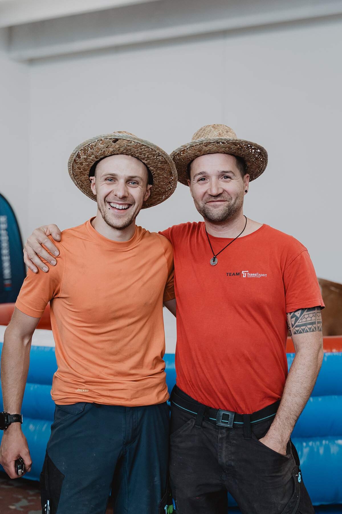 Zwei Männer stehen nebeneinander und lächeln. Beide tragen Strohhüte. Der Mann links trägt ein orangefarbenes T-Shirt, während der Mann rechts ein rotes T-Shirt trägt. Der Hintergrund ist unscharf.