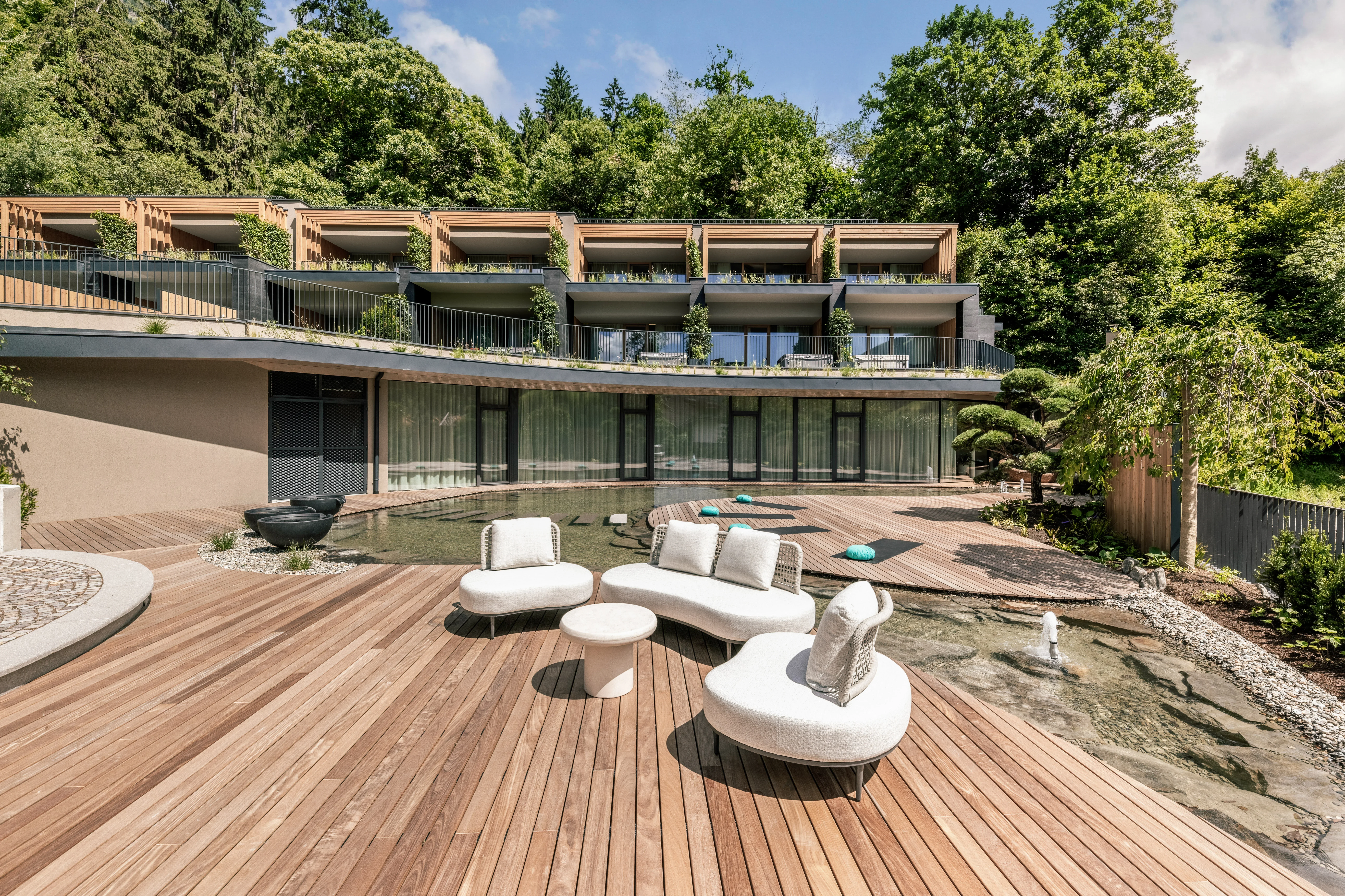 Moderne Hangvilla mit großen Fenstern, Balkonen und einer großzügigen Holzterrasse. Die Terrasse ist mit weißen Loungemöbeln ausgestattet und von grünem Wald umgeben. Ein kleiner Teich und gepflegte Gärten sind im Vordergrund zu sehen.