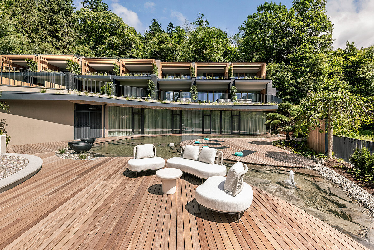 Moderne Hangvilla mit großen Fenstern, Balkonen und einer großzügigen Holzterrasse. Die Terrasse ist mit weißen Loungemöbeln ausgestattet und von grünem Wald umgeben. Ein kleiner Teich und gepflegte Gärten sind im Vordergrund zu sehen.