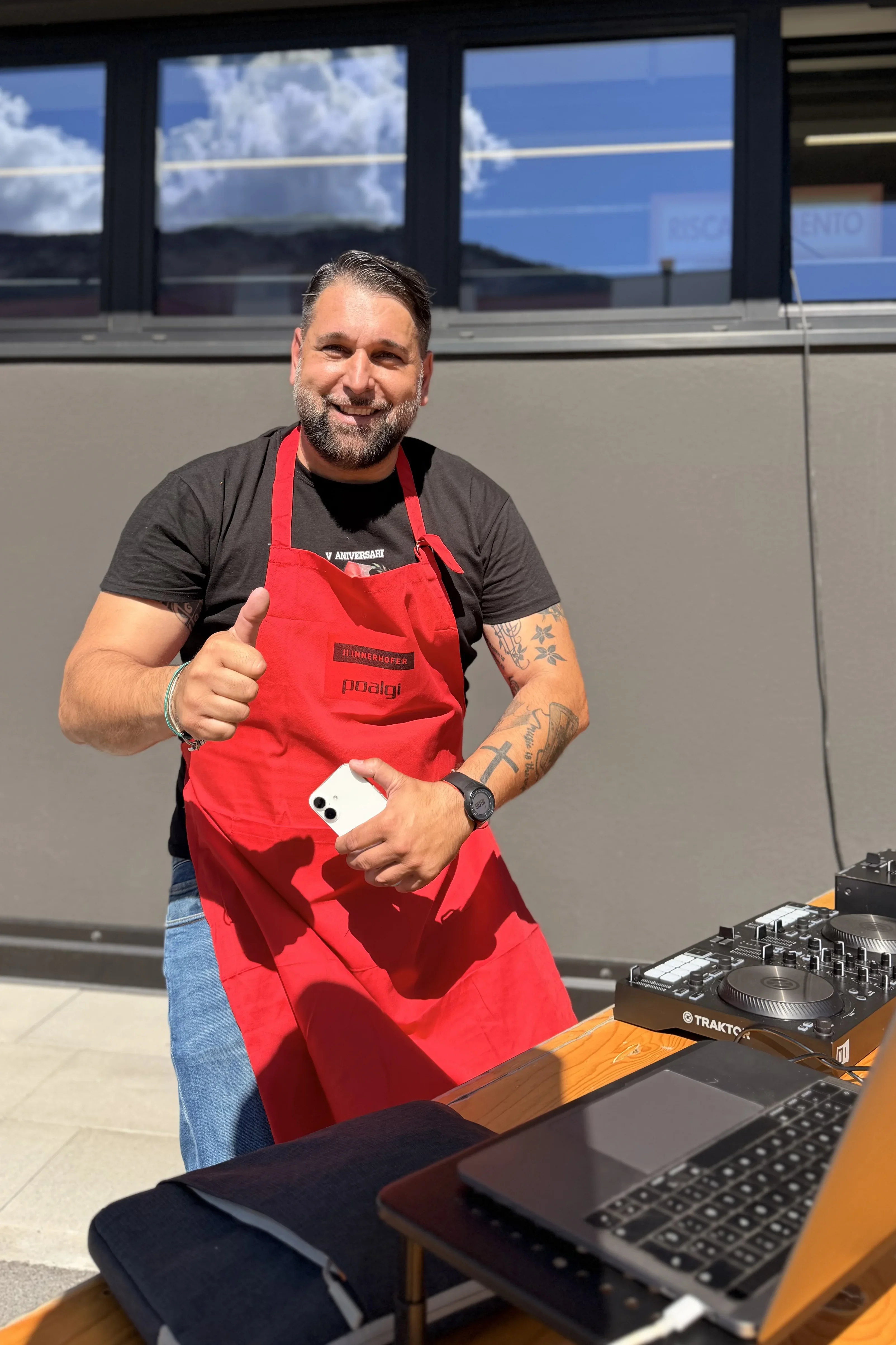 Ein Mann mit roter Schürze steht bei Sonnenschein vor einem DJ-Pult, lächelt und zeigt mit einem Daumen nach oben. Er hält ein Smartphone in der anderen Hand. Hinter ihm ist ein Gebäude mit Fenstern und ein blauer Himmel zu sehen.