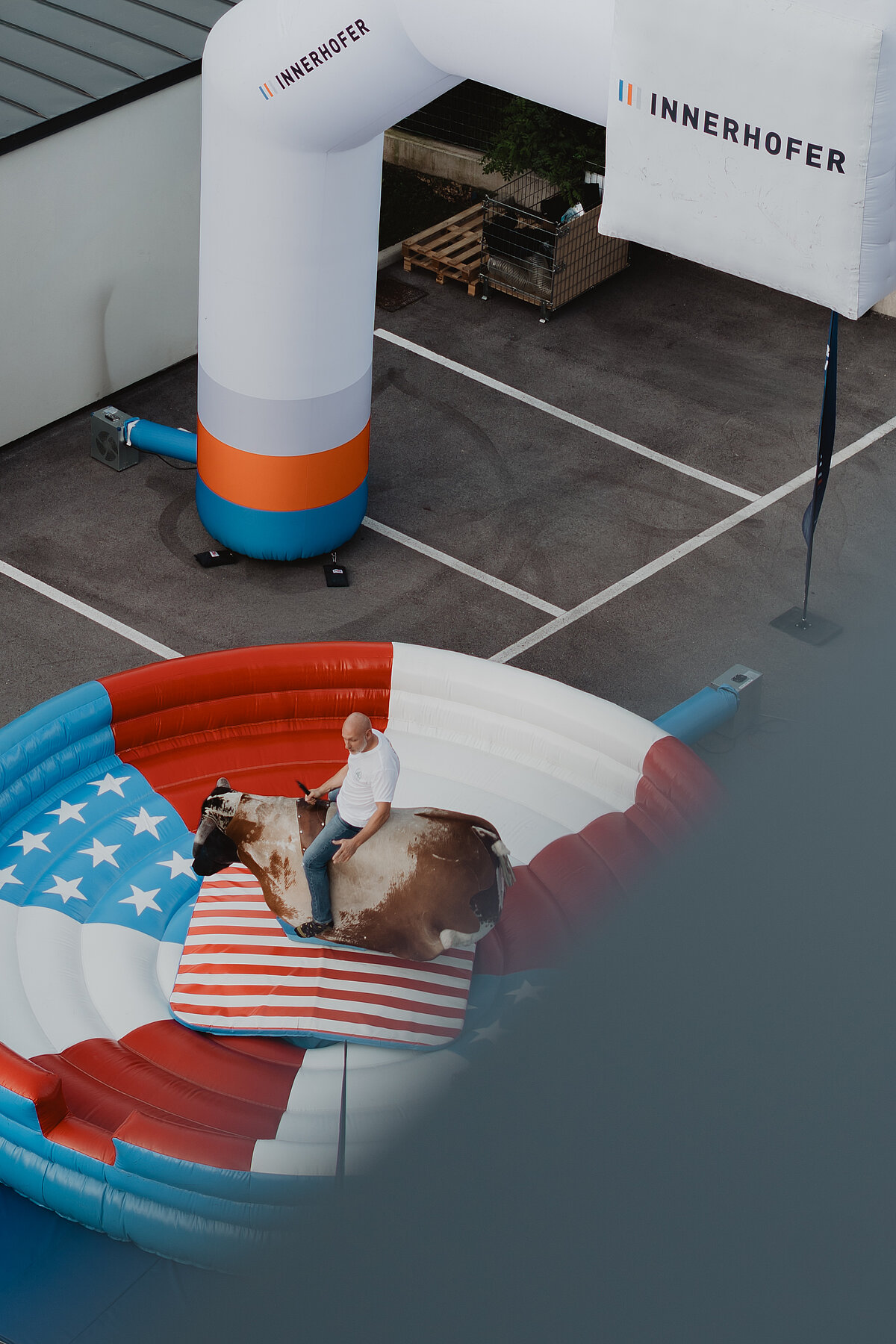Ein Mann reitet auf einem mechanischen Bullen in einem aufblasbaren Ring, der mit amerikanischen Flaggenmustern gestaltet ist. Im Hintergrund steht ein Gebläse mit dem Logo "Innerhofer".