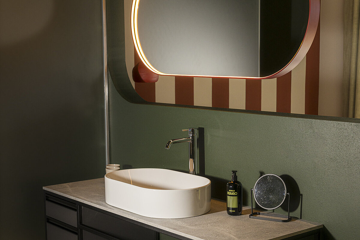 Ein modernes Badezimmer mit rechteckigem Spiegel, der von einem Licht umrahmt ist. Darunter ein Waschbecken auf einem schlichten, schwarzen Waschtisch mit Handtüchern und Seifenspender. Die Wände sind in Streifen und dunklen Tönen gestaltet.