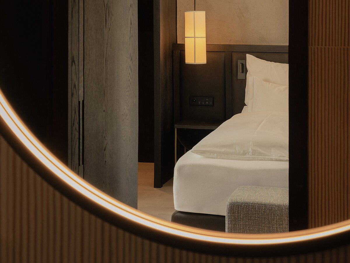 Ein modernes Hotelzimmer mit einem Doppelbett, das durch eine runde Öffnung sichtbar ist. An der Wand hängt eine beleuchtete, zylindrische Lampe. Die Einrichtung wirkt minimalistisch und elegant.