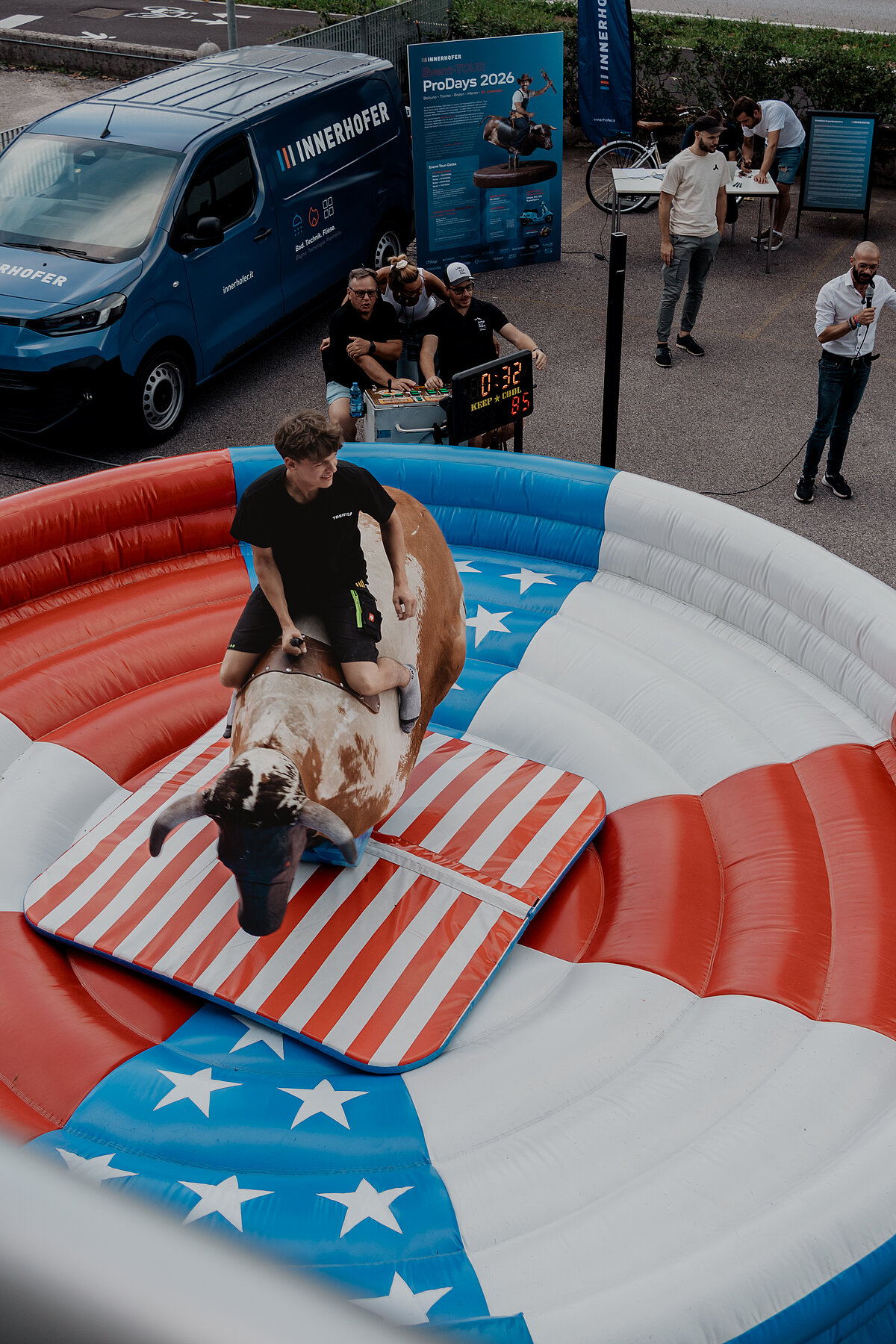 Ein Mann reitet auf einem mechanischen Bullen in einer aufblasbaren Rodeo-Arena mit amerikanischer Flaggenoptik. Im Hintergrund bedienen Personen die Steuerung des Bullen. Eine digitale Anzeige zeigt die benötigte Zeit. Links steht ein blauer Transporter mit offener Tür.