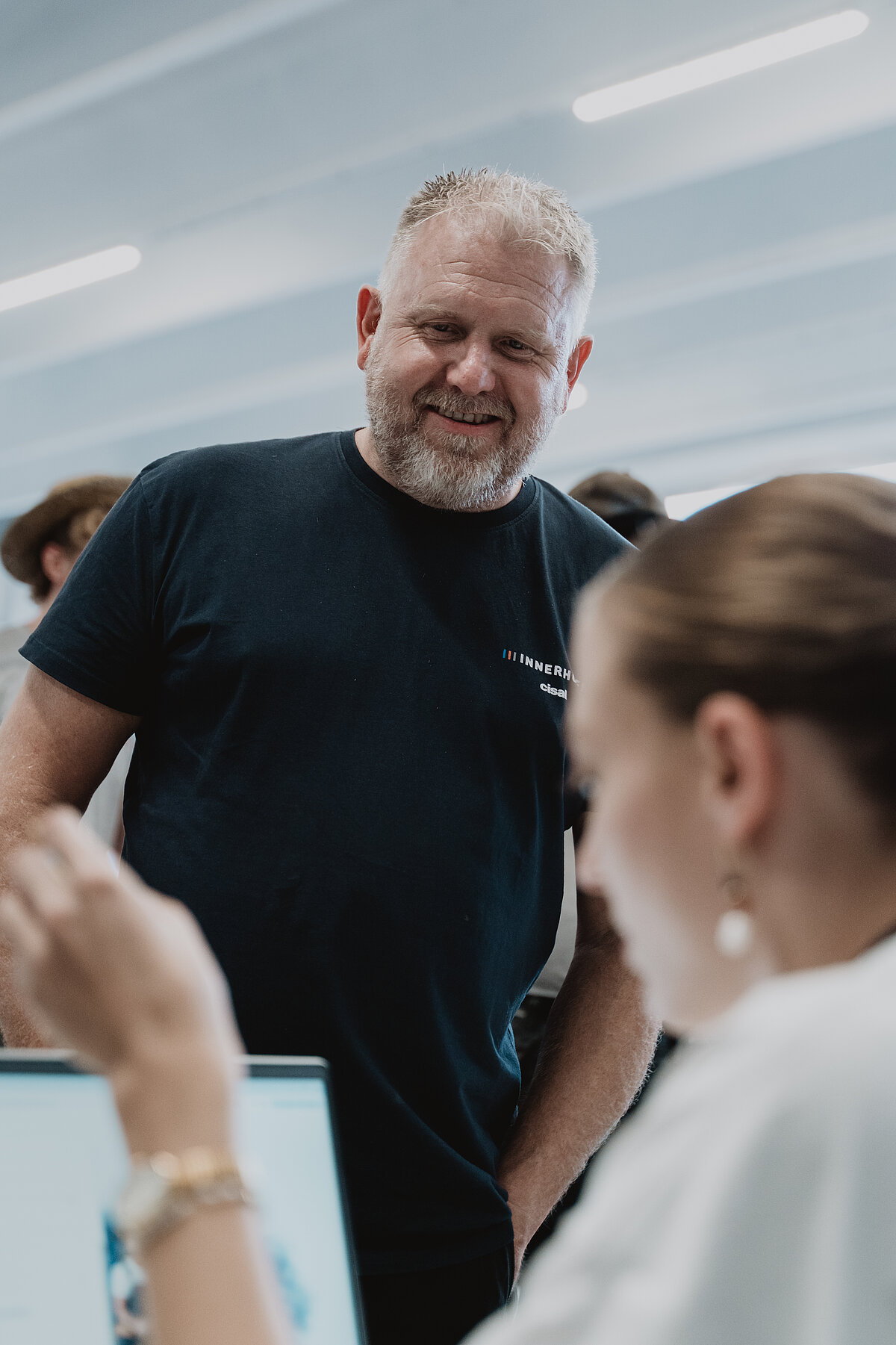 Ein Mann mit grauem Bart und schwarzem T-Shirt lächelt und steht in einem modernen Büro. Eine Frau im Vordergrund arbeitet an einem Laptop. Die Atmosphäre wirkt professionell und freundlich.