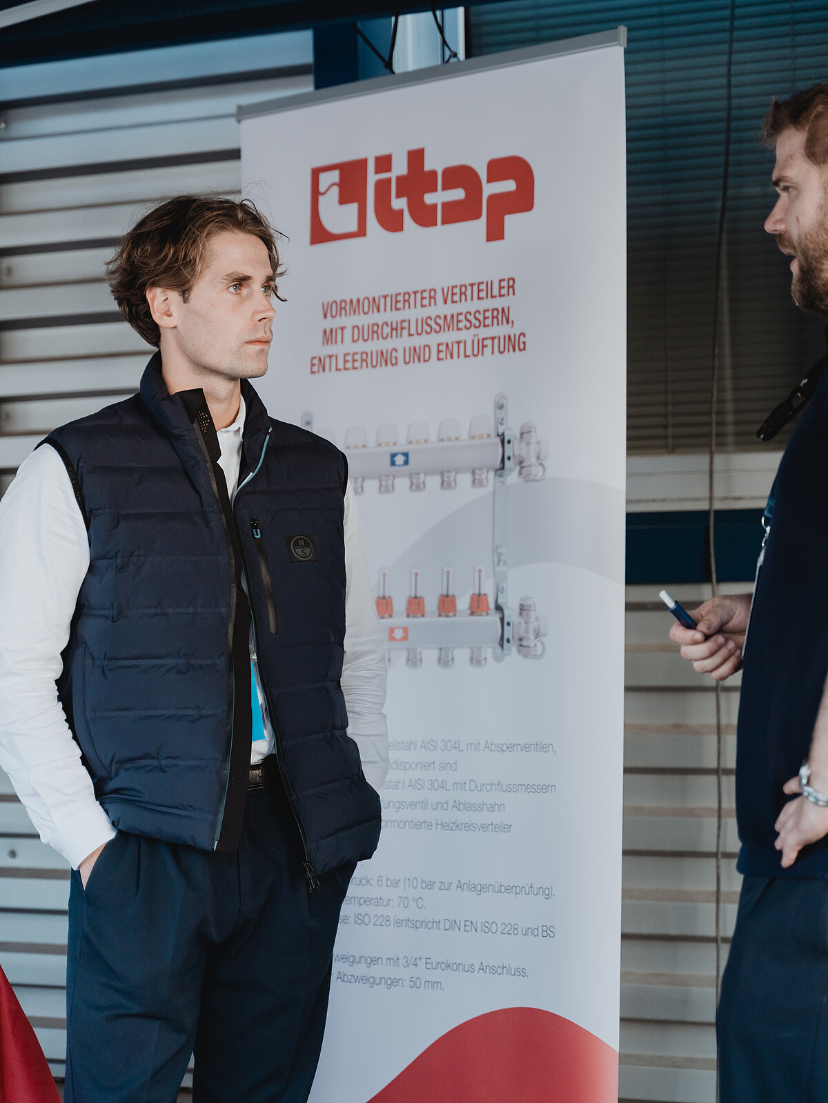 Zwei Männer unterhalten sich vor einem Werbebanner der Firma "Itap". Einer der Männer hält ein Mobiltelefon in der Hand. Das Banner beschreibt vorinstallierte Verteiler mit Durchflussmessern und weiteren Funktionen. Die Männer tragen lässige Kleidung.