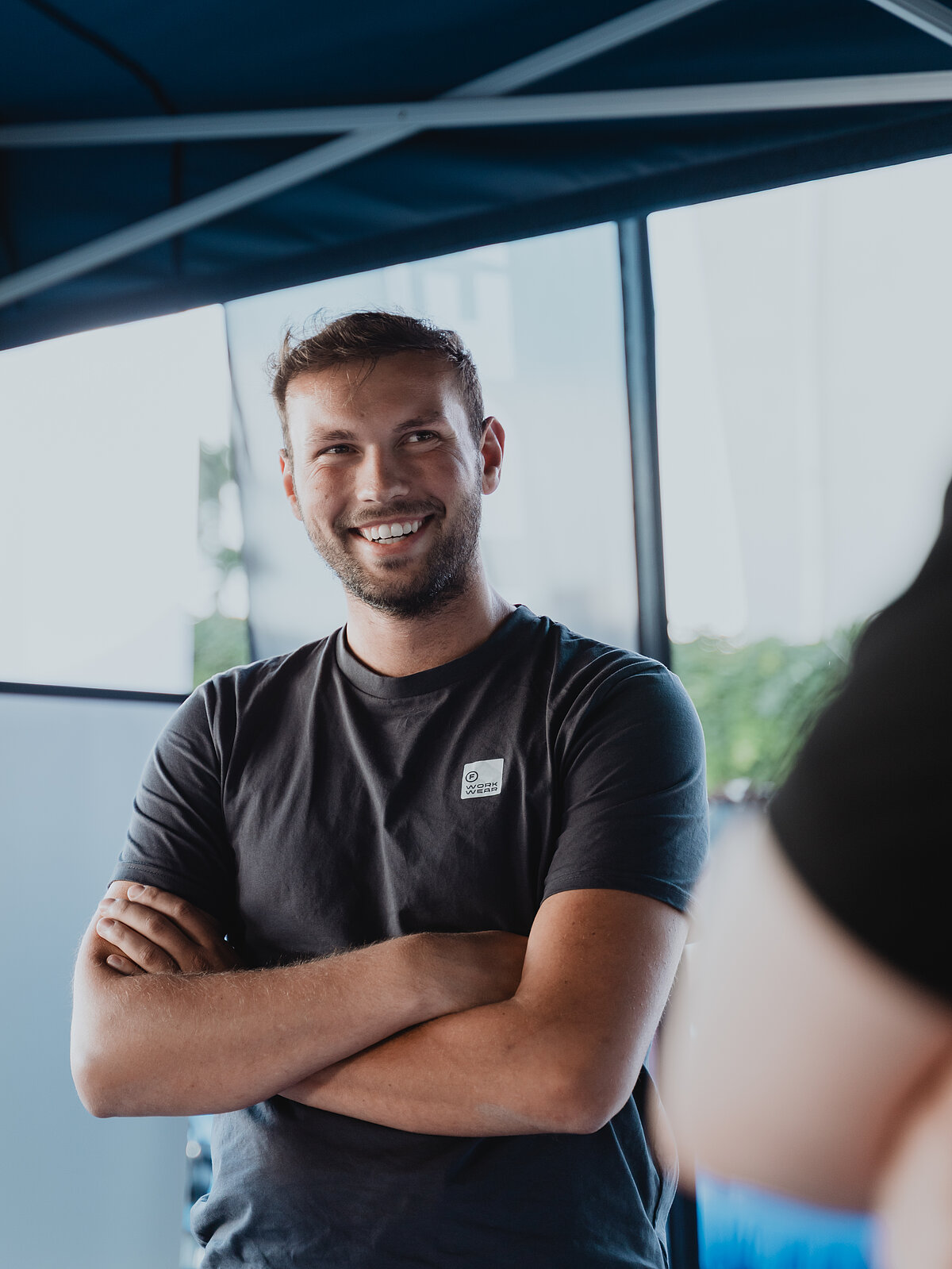 Ein lächelnder Mann mit Bart steht mit verschränkten Armen unter einem blauen Pavillon. Er trägt ein dunkles T-Shirt und wirkt entspannt und freundlich. Im Hintergrund sind unscharf Bäume und ein Stück Himmel zu erkennen.