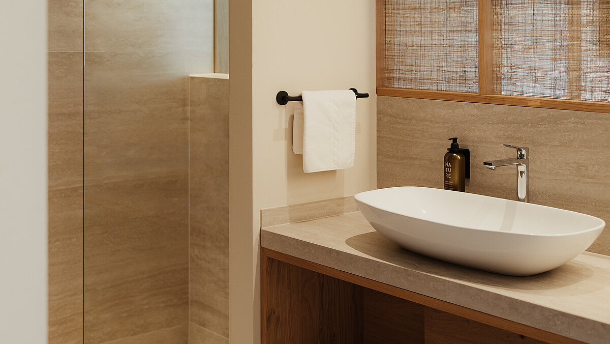 Modernes Badezimmer mit begehbarer Dusche, Regenbrause, Waschbecken mit Wasserhahn und Seifenspender. Ein Holzregal darunter hält saubere Handtücher. Große Fenster mit Holzjalousien sorgen für natürliches Licht. Neutrale Farbtöne verleihen dem Raum eine entspannende Atmosphäre.