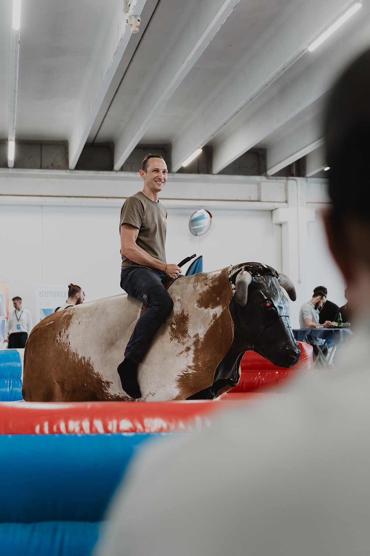 Eine Person reitet auf einem mechanischen Bullen in einem Indoor-Bereich. Im Vordergrund befinden sich verschwommene Köpfe von Zuschauern, die das Geschehen beobachten. Der Raum hat eine moderne Decke mit Beleuchtung.