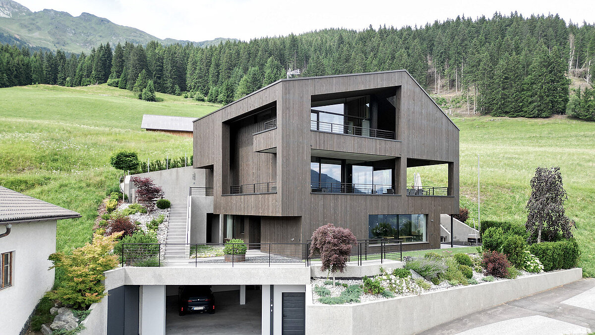 Modernes Holzhaus auf einem Hügel mit großer Terrasse und Garten. Die Architektur ist kantig und kubistisch. Im Vordergrund eine offene Garage. Die umliegende Landschaft ist grün, mit einem Hintergrund aus Wäldern und Bergen. Ein idyllischer, zeitgemäßer Wohnsitz in ländlicher Umgebung.