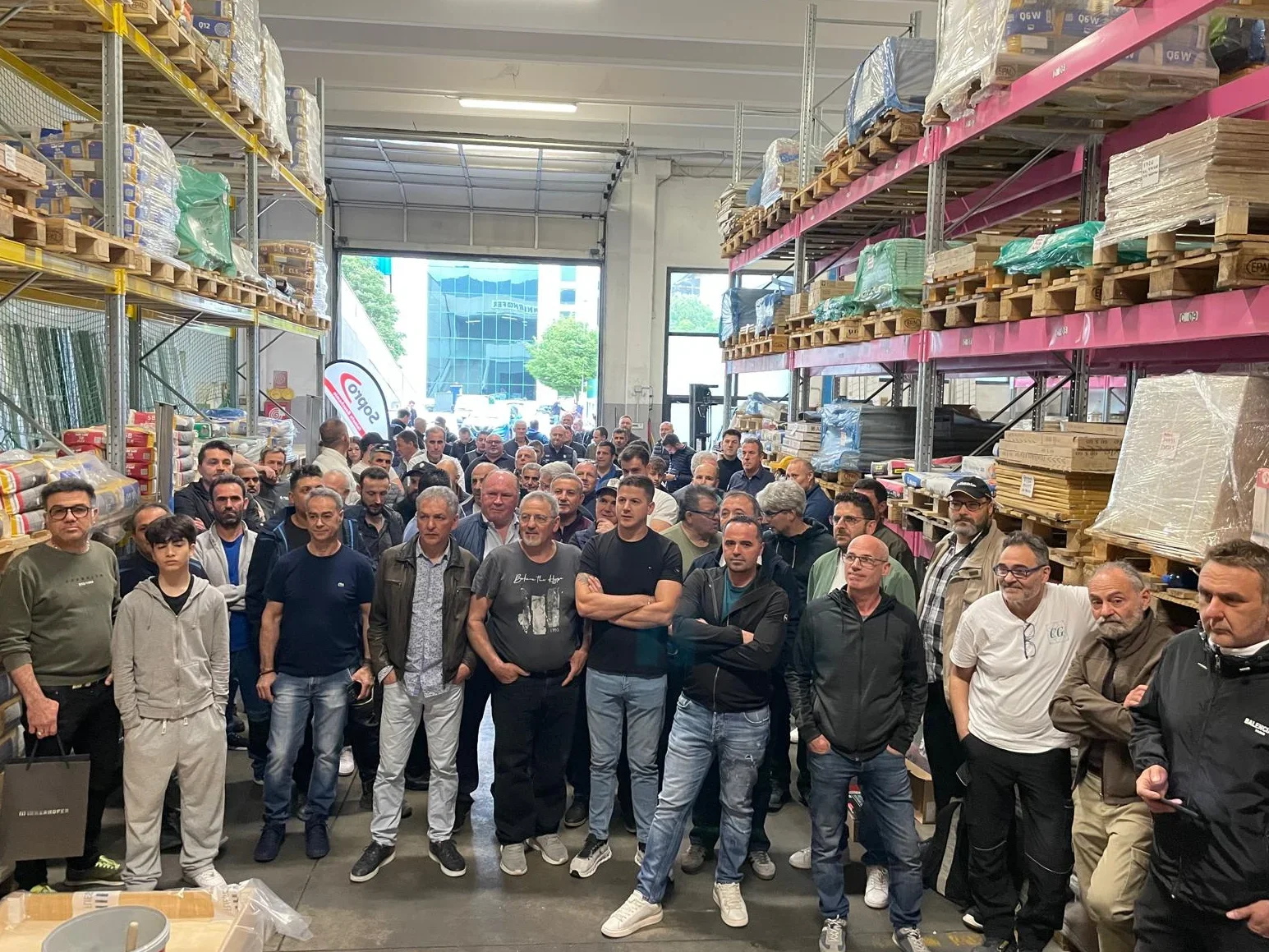Eine große Gruppe von Menschen versammelt sich in einem Lagerhaus mit Regalen voller Material. Sie schauen alle in dieselbe Richtung, als ob sie jemandem außerhalb des Bildes zuhören oder zusehen würden. Die Umgebung ist industriell und funktional gestaltet.