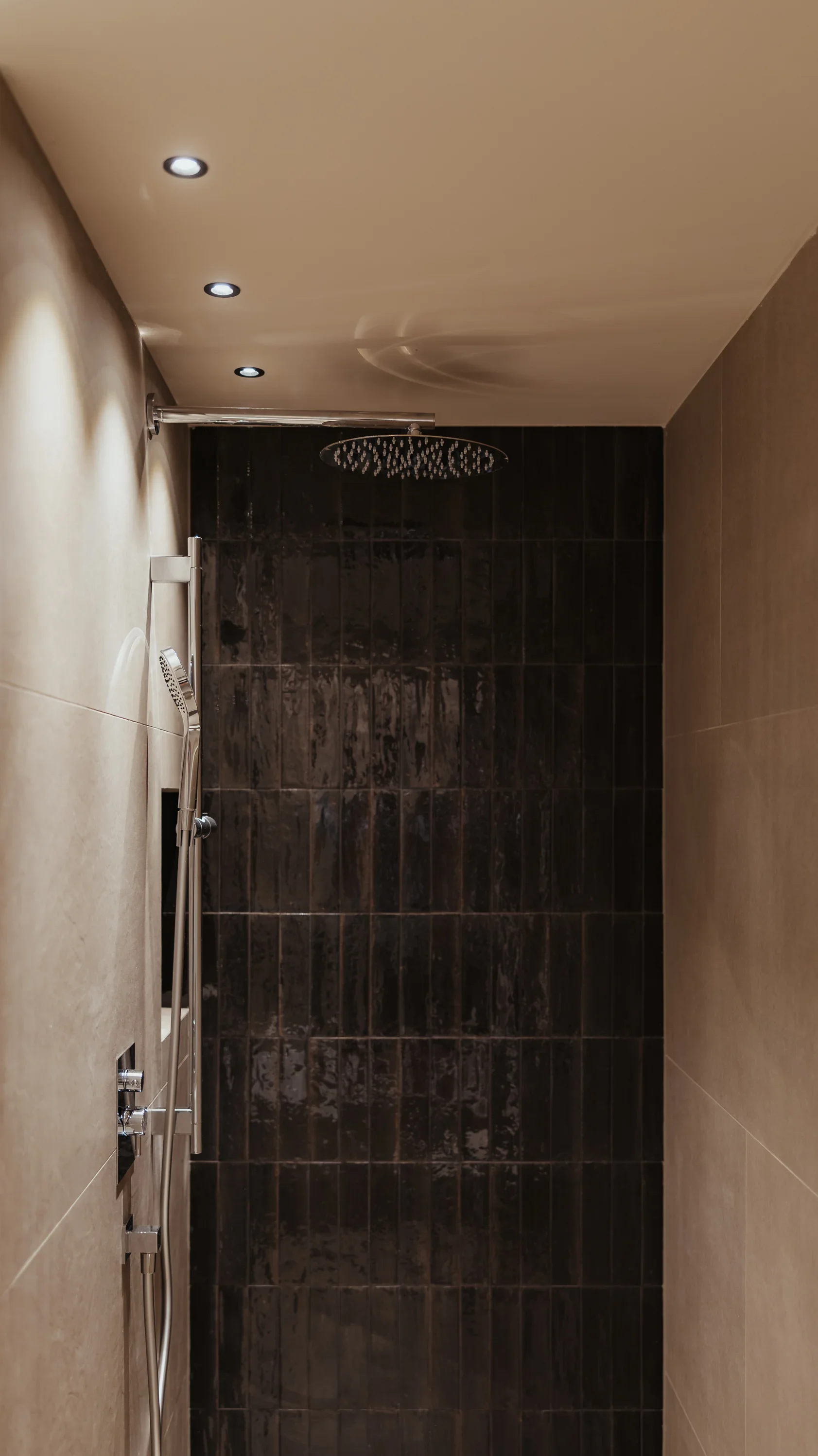 Moderne Dusche mit dunkel gefliester Wand, Regenduschkopf und seitlicher Handbrause. Deckenbeleuchtung mit drei Einbauspots sorgt für dezente Ausleuchtung. Wände in neutralem Beige.