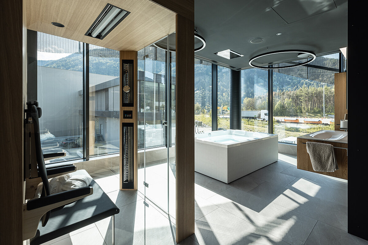 Moderner Wellnessbereich mit großer Glassauna, freistehender Badewanne und hellem Tageslicht. Durch die großen Fenster blickt man auf eine bergige Landschaft. Hochwertige Materialien und minimalistisches Design prägen den Raum.