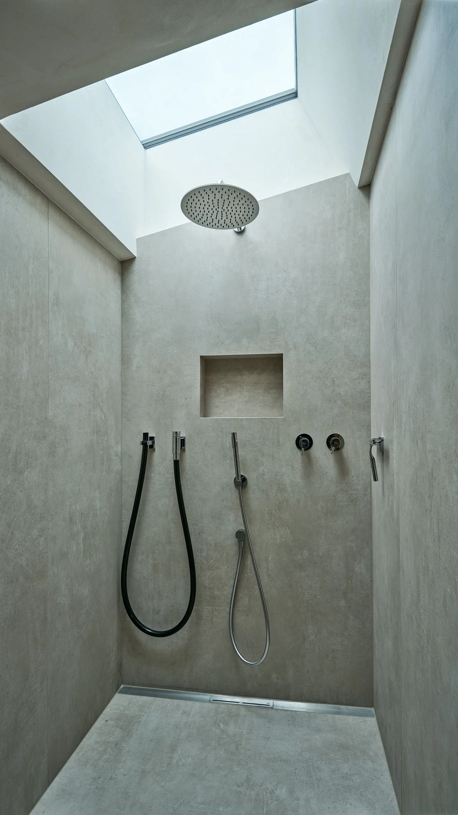 Moderne, minimalistische Dusche mit grauer Betonoptik, großer Regendusche an der Decke und zwei schwarzen Handbrausen. Ein rechteckiges Wandregal ist in der Duschwand eingelassen. Oberhalb ein Dachfenster, das natürliches Licht hereinlässt.