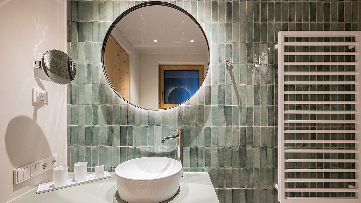 Ein modernes Badezimmer mit rundem Spiegel über einem weißen Waschbecken. Die grüne Wand besteht aus vertikalen Fliesen. Rechts neben dem Waschbecken befindet sich ein weißer Handtuchheizkörper. Dezente Beleuchtung schafft eine entspannte Atmosphäre.