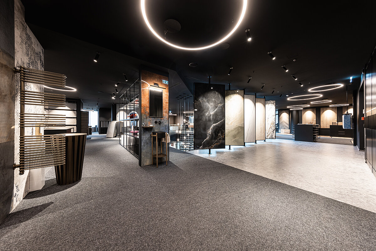Innenansicht eines modernen Ausstellungsraums mit Teppichboden, eleganter Beleuchtung und verschiedenen Fliesenmustern an den Wänden. Eine Mischung aus Stein- und Parkettoptik ist deutlich sichtbar. Die Atmosphäre wirkt ruhig und stilvoll.