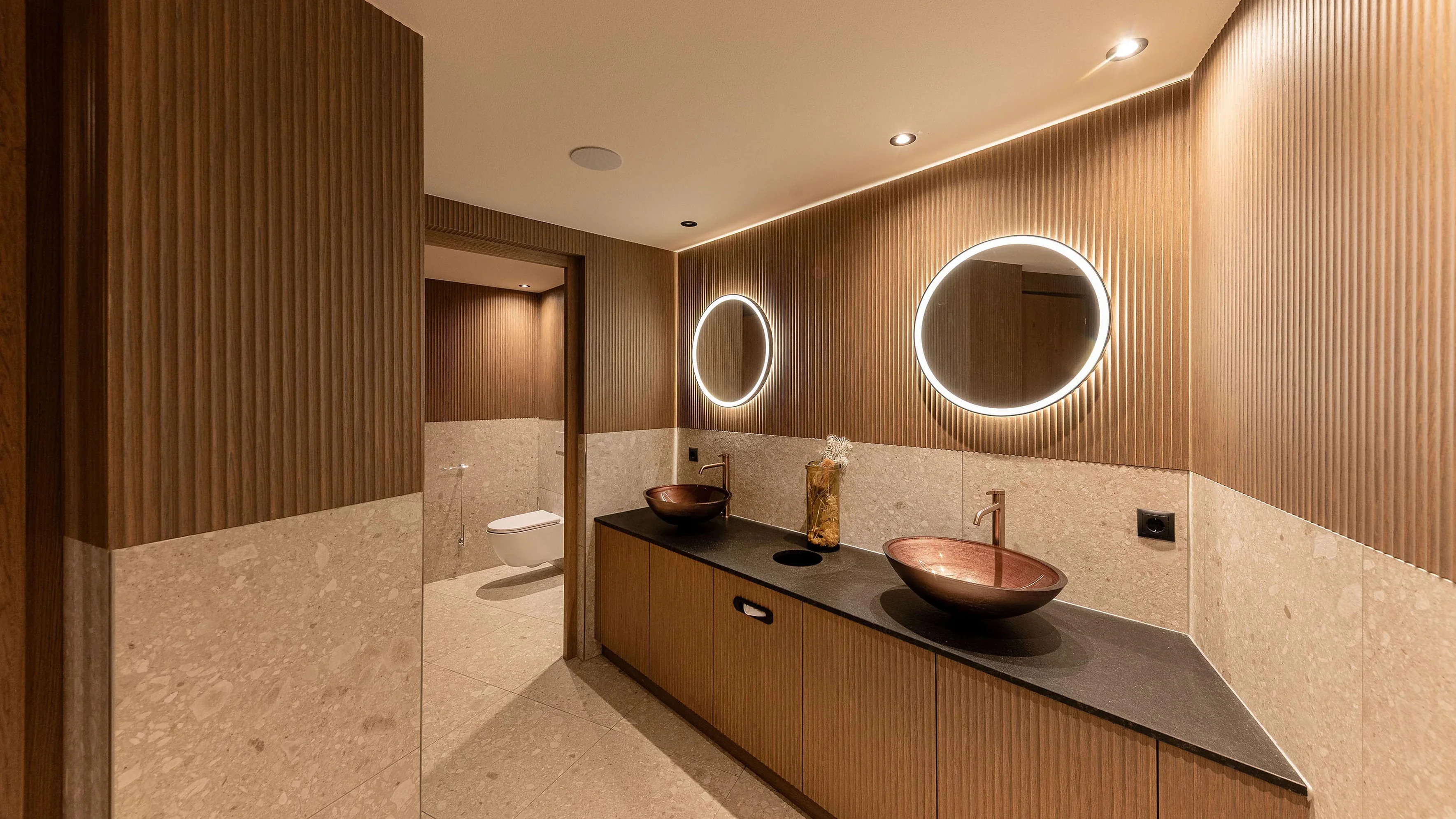 Modernes Badezimmer mit zwei runden Spiegeln und Waschbecken aus Kupfer. Holzverkleidung und dezente Beleuchtung verleihen dem Raum eine warme Atmosphäre. Links ist ein separater Bereich mit einer Toilette sichtbar.