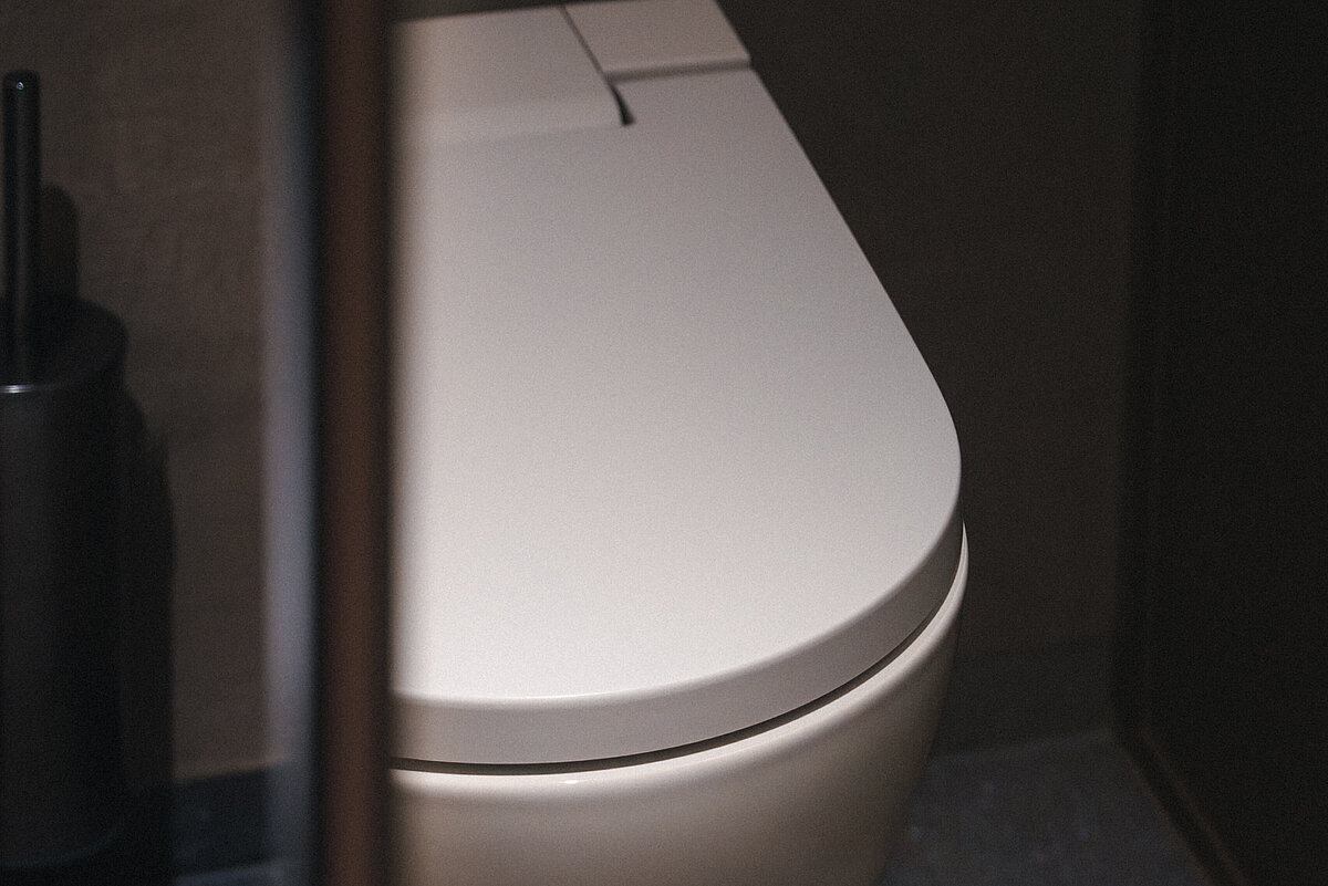 Modernes, weißes Wand-WC in einem gedämpft beleuchteten Badezimmer mit dunklen, minimalistischen Wänden und Boden. Links steht eine Toilettenbürste in einem Halter.