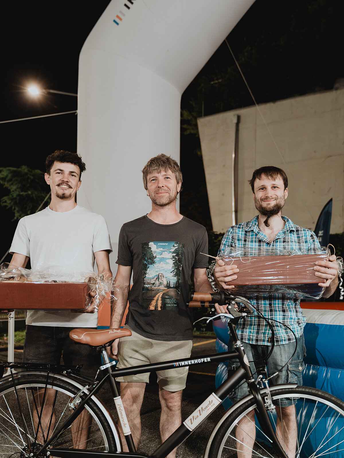 Drei Männer posieren vor einem aufblasbaren Torbogen. Zwei von ihnen halten verpackte Geschenke, während ein Fahrrad im Vordergrund steht. Es ist Nacht, erkennbar durch die Beleuchtung im Hintergrund. Alle drei wirken entspannt und lächeln leicht.