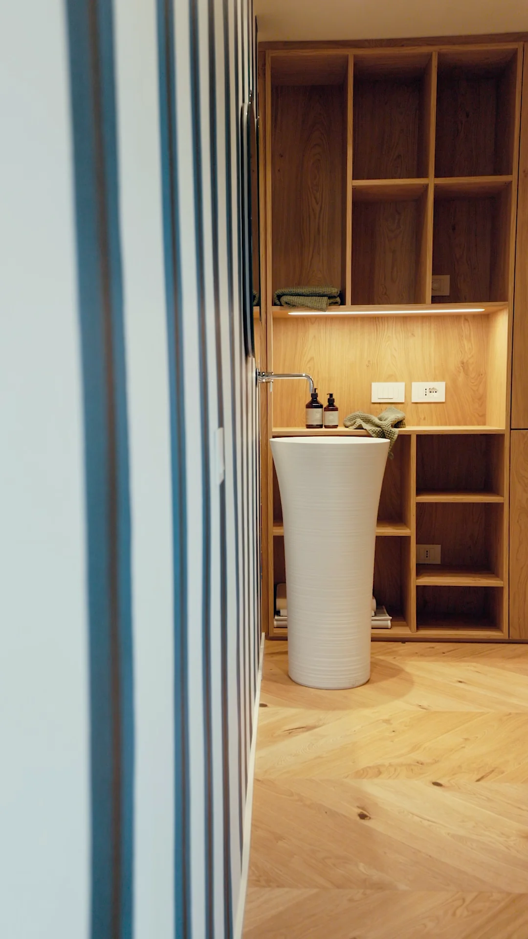 Ansicht eines modernen Badezimmers mit einem schmalen, hohen Waschbecken und einer Wand aus offenen Holzregalen. Zwei Flaschen Pflegeprodukte stehen neben dem Wasserhahn. Der Boden ist aus Holz, und die Wand ist gestreift.