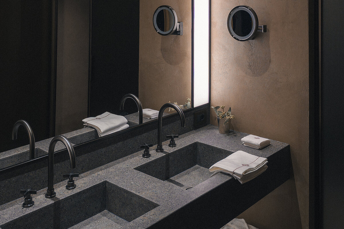 Modernes Badezimmer mit Doppelwaschbecken aus Stein, zwei schwarzen Wasserhähnen und integriertem Spiegel mit beleuchtetem Rahmen. Darunter ein Regal mit weißen Handtüchern. Dezente Wandbeleuchtung schafft eine elegante, ruhige Atmosphäre.