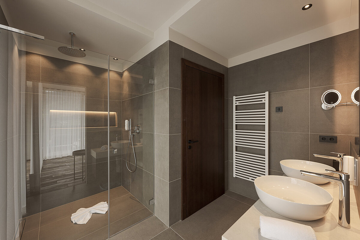 Modernes Badezimmer mit Regendusche in einer Glasdusche, zwei weißen Waschbecken auf einem eleganten Waschtisch, beleuchtetem Spiegel und Handtuchwärmer. Graue Wand- und Bodenfliesen schaffen eine stilvolle und entspannende Atmosphäre.