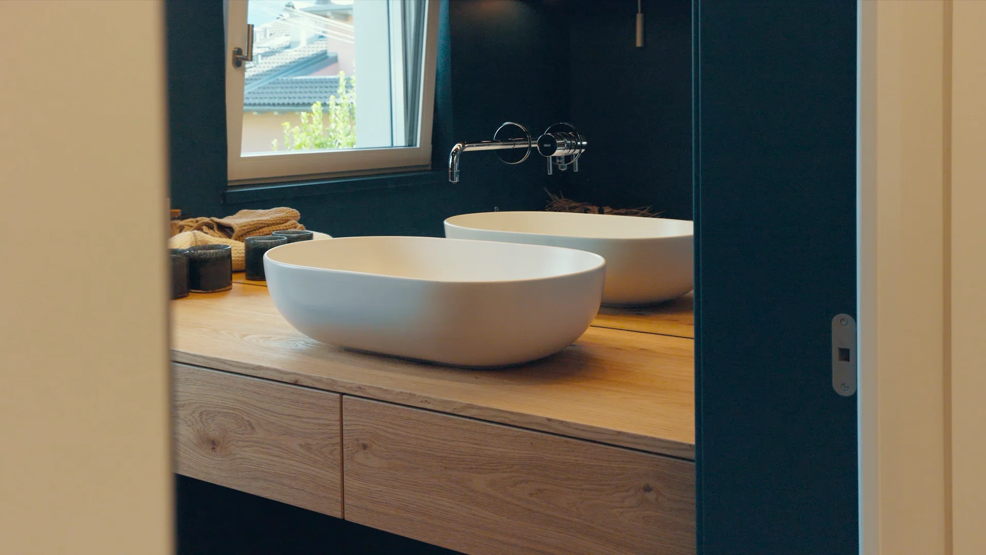 Ein modernes Badezimmer mit einem ovalen, weißen Waschbecken auf einer hölzernen Arbeitsplatte. Im Hintergrund ist ein Fenster und eine Wandarmatur zu sehen. Tageslicht fällt durch das Fenster und beleuchtet den Raum.