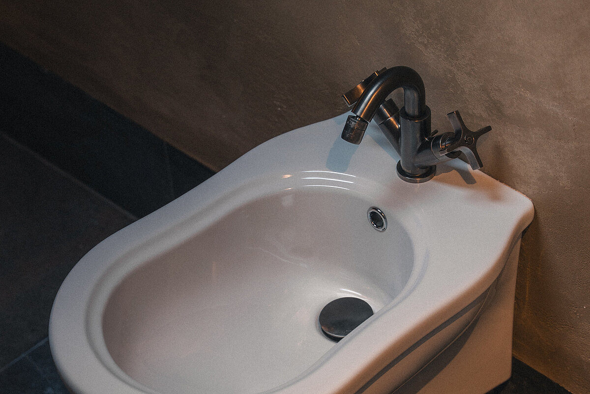 Ein Bidet mit einem Wasserhahn und einem Metallgriff in einem Badezimmer. Der Boden um das Bidet ist aus dunklem Stein. Das Bidet ist an einer taupefarbenen Wand montiert.