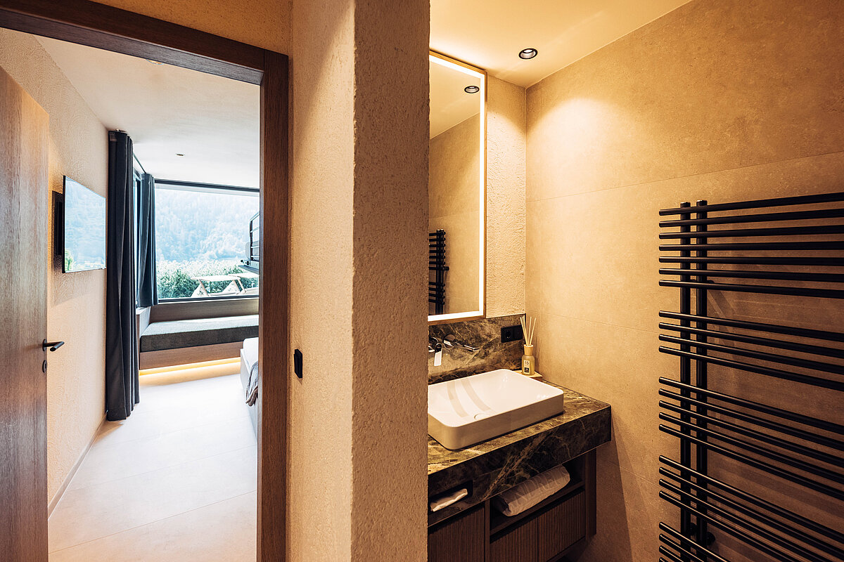 Blick vom Badezimmer in ein Schlafzimmer mit großem Fenster. Im Badezimmer sind ein moderner Waschtisch mit Spiegel und ein Handtuchheizkörper zu sehen. Das Schlafzimmer hat einen hellen Boden und bietet Aussicht auf eine Landschaft.