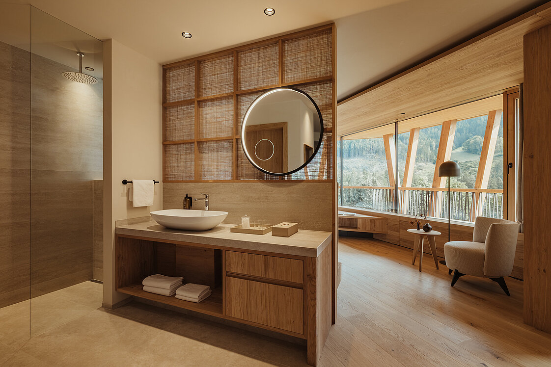 Modernes Badezimmer mit Holzverkleidung, freistehendem Waschbecken und rundem Spiegel. Rechts befindet sich ein Wohnbereich mit Sesseln und Panoramafenstern, die einen Blick auf die Natur bieten. Angenehmes, warmes Licht durchflutet den Raum und schafft eine entspannende Atmosphäre.
