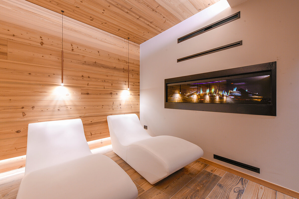 Zwei moderne, weiße Liegestühle in einem stilvollen Raum mit Holzwänden. An der Wand befindet sich ein elektrischer Kamin. Die Beleuchtung ist warm und dezent. Die Atmosphäre wirkt entspannt und einladend.