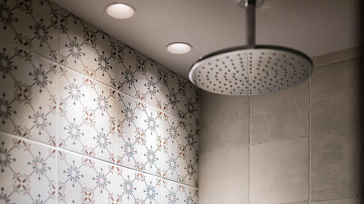 Moderne Dusche mit zwei verschiedenen Fliesendesigns: links florale Muster, rechts schlichte, helle Quadrate. Eine große, runde Regendusche hängt an der Decke, darunter eine Handbrause und ein integriertes Fach für Duschprodukte. Deckenlichter beleuchten die Dusche.