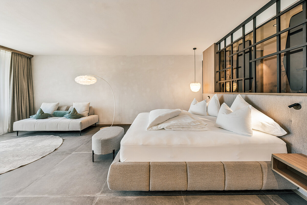 Ein modernes Schlafzimmer mit neutralen Tönen. Ein großes Bett mit weißen Kissen und Decken steht im Vordergrund, daneben ein kleiner, ovaler Hocker. Im Hintergrund befindet sich eine gemütliche Sitzecke mit Kissen. Eine große Fensterfront mit Vorhängen sorgt für Helligkeit und Offenheit.