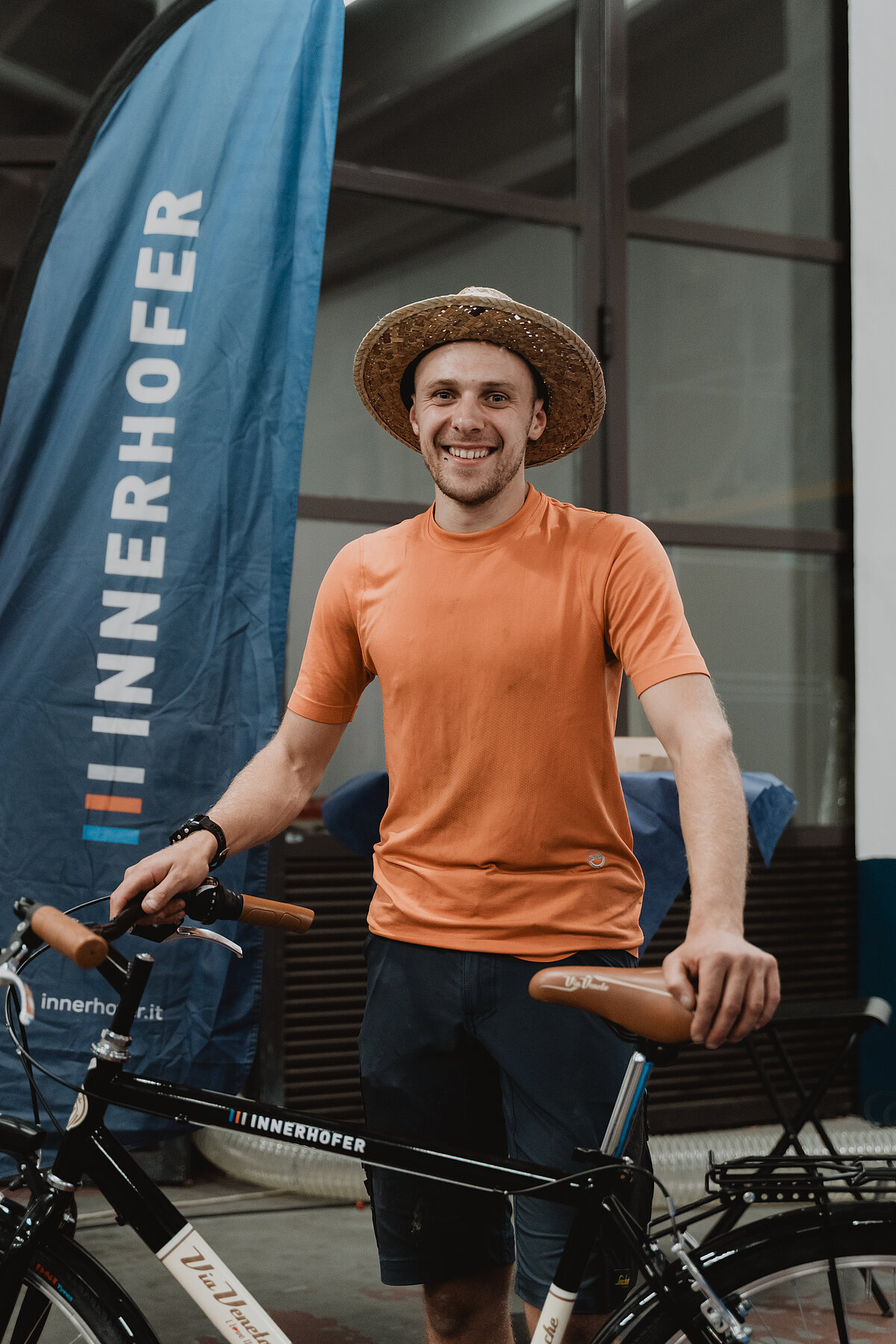 Ein Mann steht lächelnd neben einem Fahrrad, trägt ein orangefarbenes T-Shirt und einen Strohhut. Im Hintergrund ist ein blaues Banner mit der Aufschrift "INNERHOFER" sichtbar. Die Szene scheint in einem Innenraum stattgefunden zu haben.