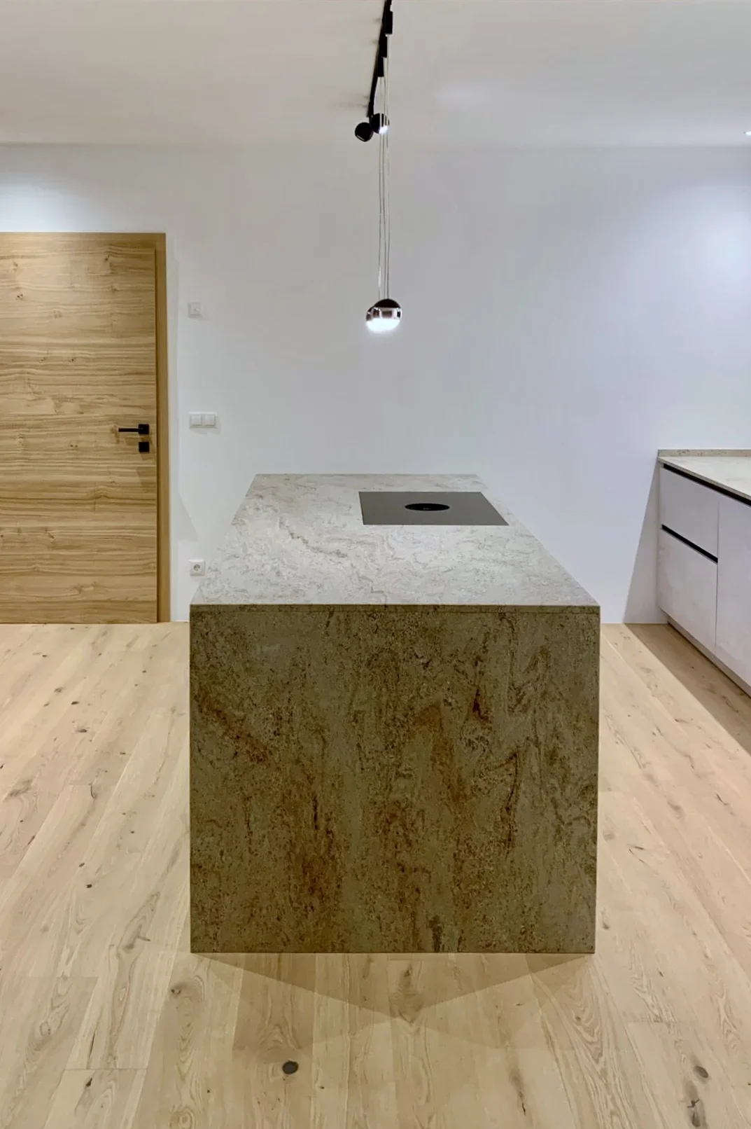 Moderne Küche mit Holzfußboden, zentraler Kochinsel aus hellem Stein mit eingebautem Herd. Weiße Wände und minimalistisches Design. Rechts befinden sich eine Spüle und dunkle Schränke. Eine schlichte Hängeleuchte beleuchtet die Insel. Holztür in der Wand links.