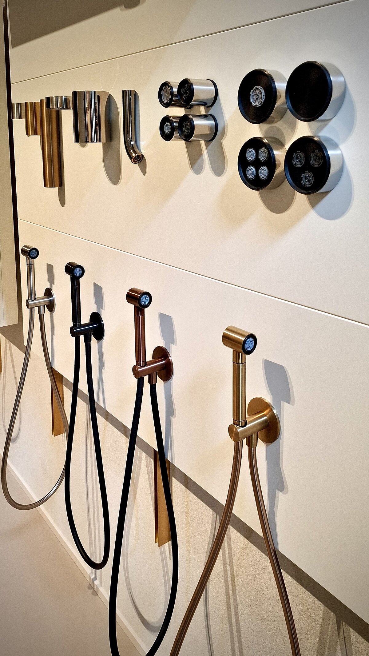 Mehrere Duschschläuche in verschiedenen Farben und Designs sind an einer Wand ausgestellt. Darüber sind passende Duschköpfe und Armaturen befestigt. Das Arrangement zeigt eine Vielfalt an Stiloptionen für Badezimmerausstattungen.