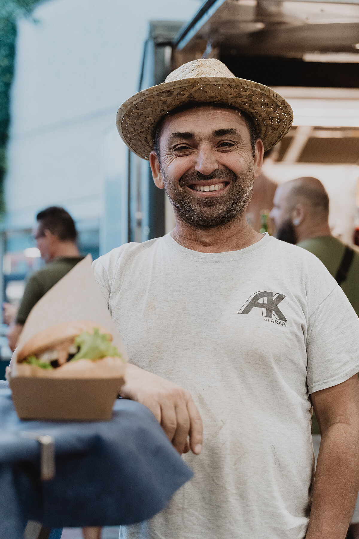 Ein Mann in einem weißen T-Shirt und Strohhut steht vor einem Imbisswagen. Er lächelt und hat einen Karton mit einem Burger in der Hand. Im Hintergrund sind unscharf weitere Personen zu erkennen.