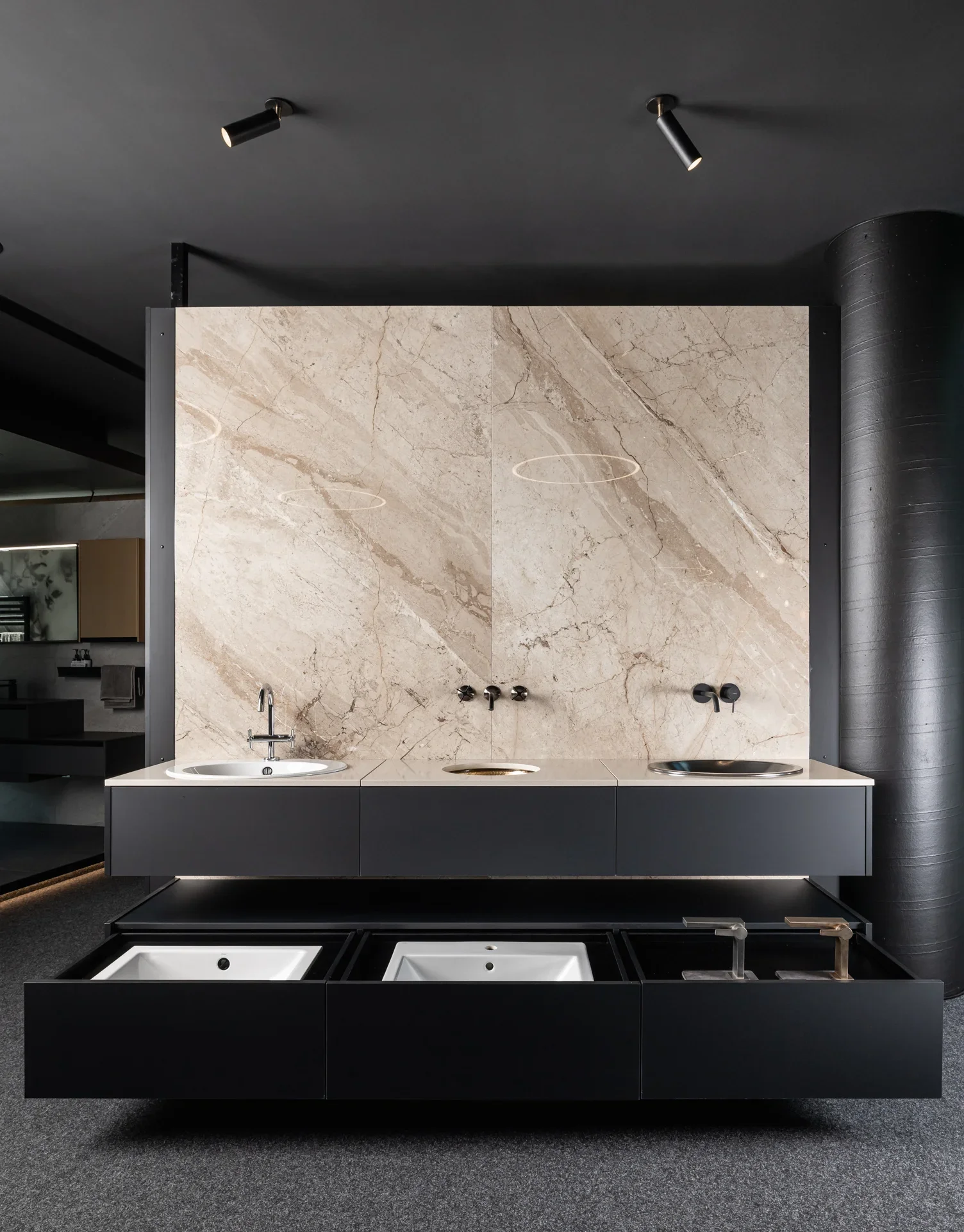 Ein modernes Badezimmer mit einer Wand aus beigem Marmor, zwei Waschbecken und zwei offenen schwarzen Schubladen darunter. Oben sind zwei Wasserhähne und eine dezente Beleuchtung von der Decke.