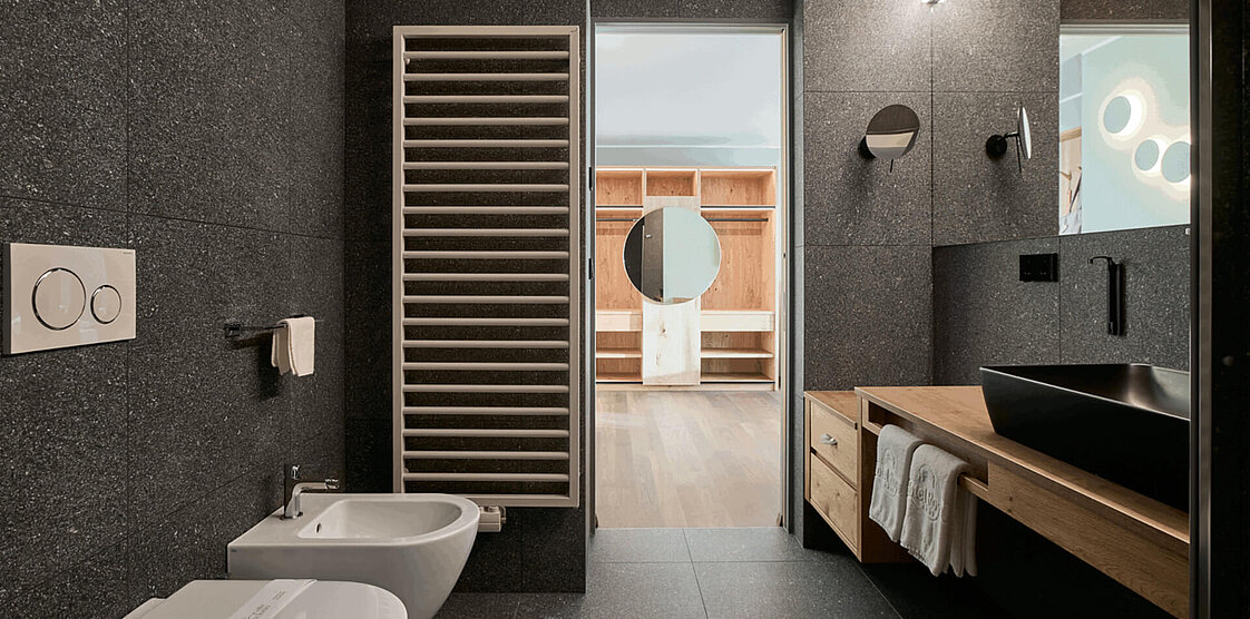 Modernes Badezimmer mit dunkler Steinwand, großem Spiegel, Holzschränken und Badewanne. Im Hintergrund ist ein Flur mit Tageslicht zu sehen.